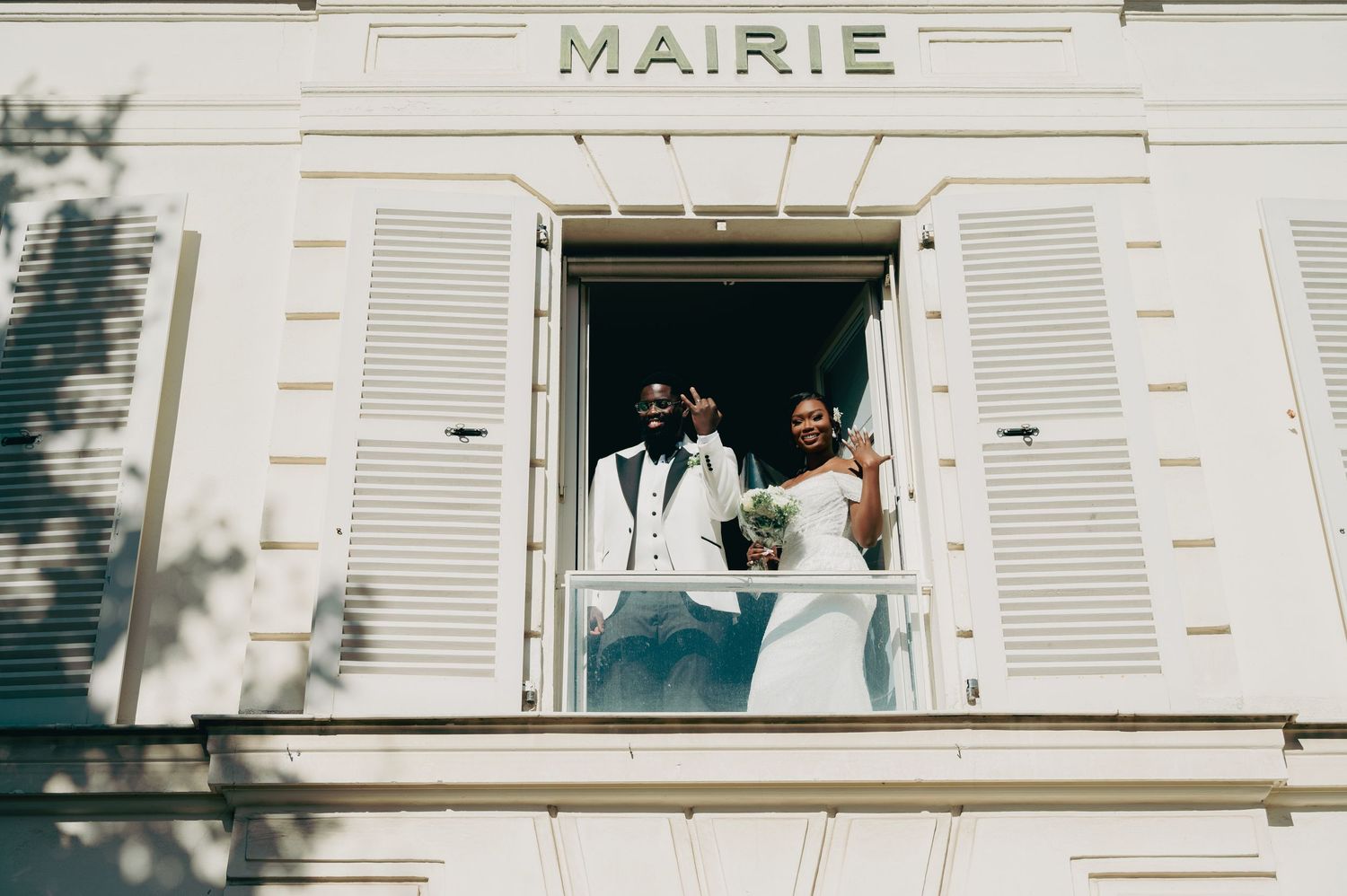 Newlywed couple waves from a balcony window of a white French Mairie building after their ceremony.