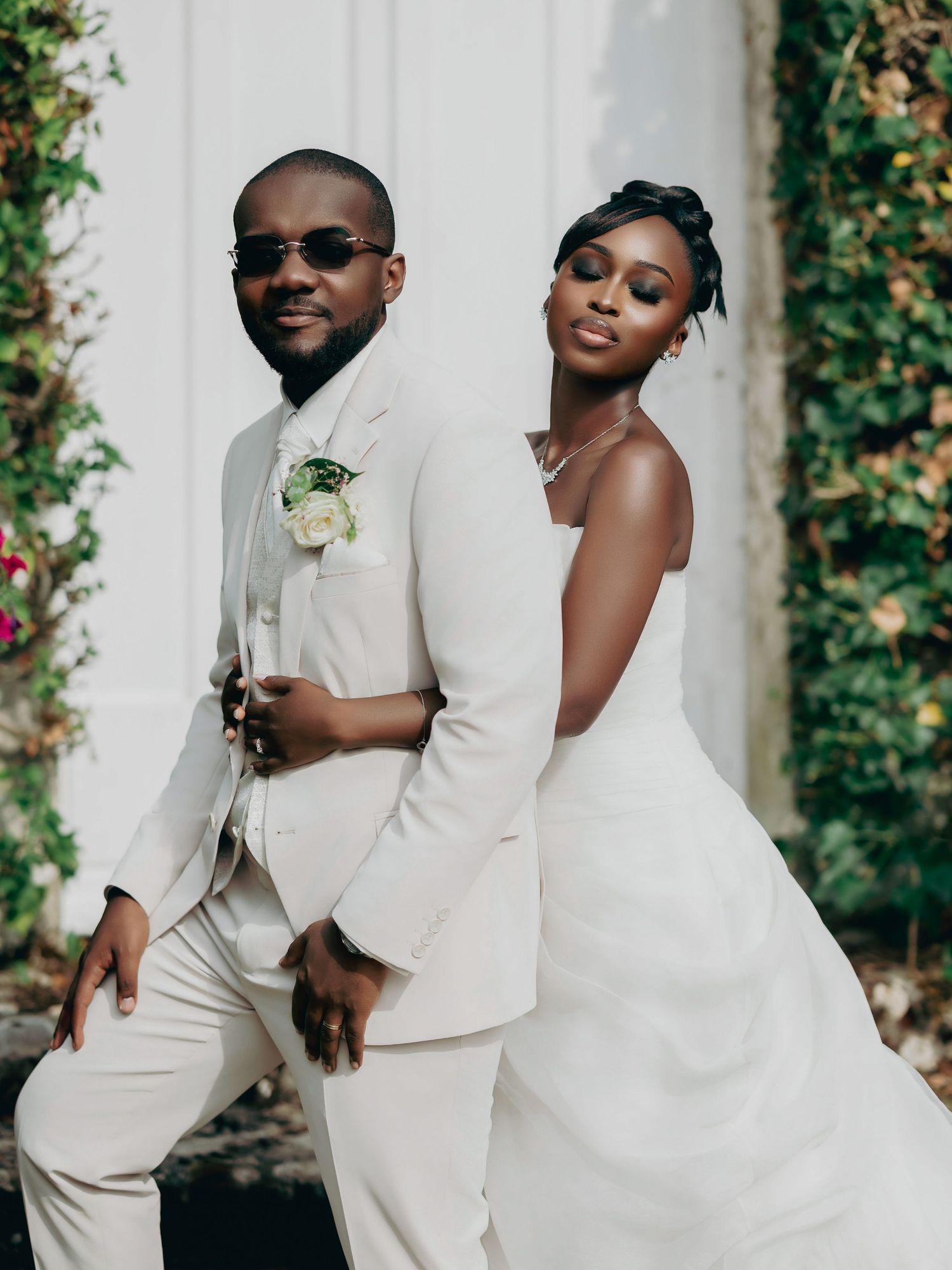 Stylish bride and groom pose back-to-back before white door, both wearing coordinating ivory outfits.