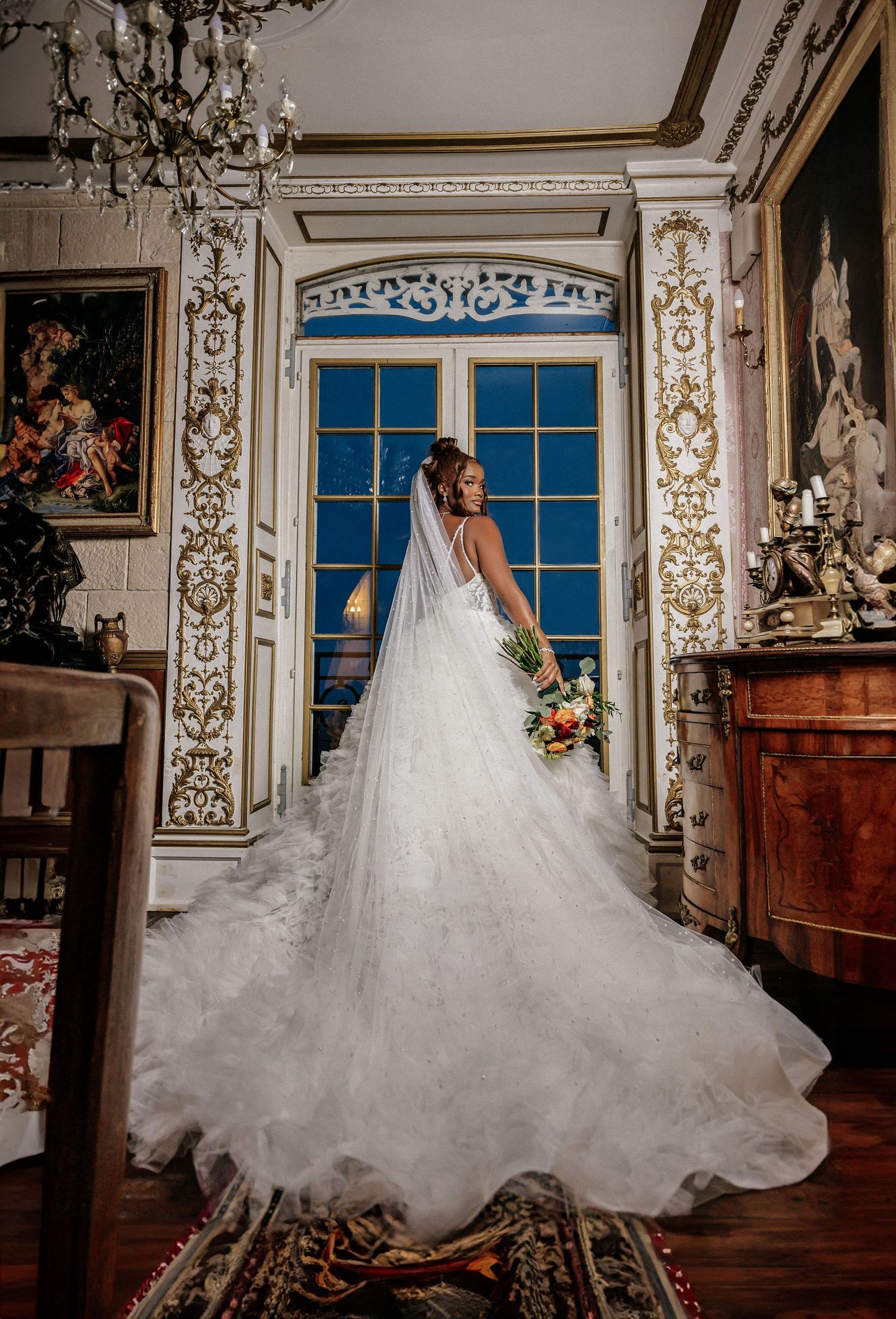 Bride in a stunning white ball gown with veil stands before ornate gold-trimmed windows in an elegant baroque room.