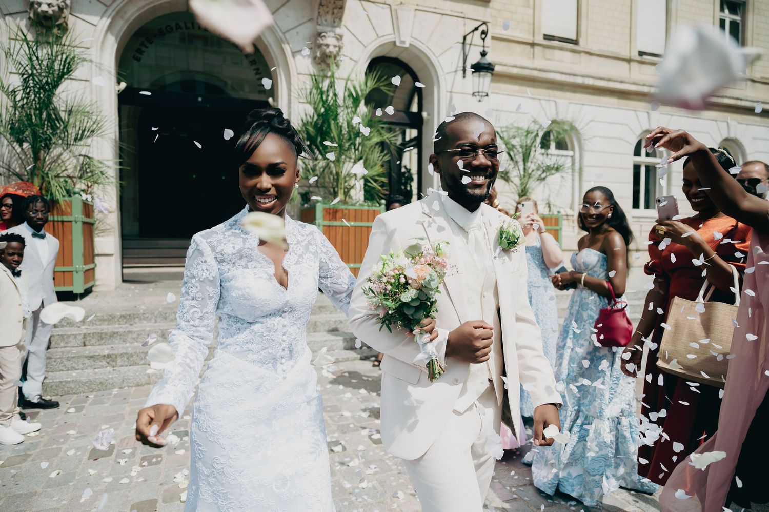Newlyweds walk joyfully through confetti shower outside elegant building, bride in lace gown.