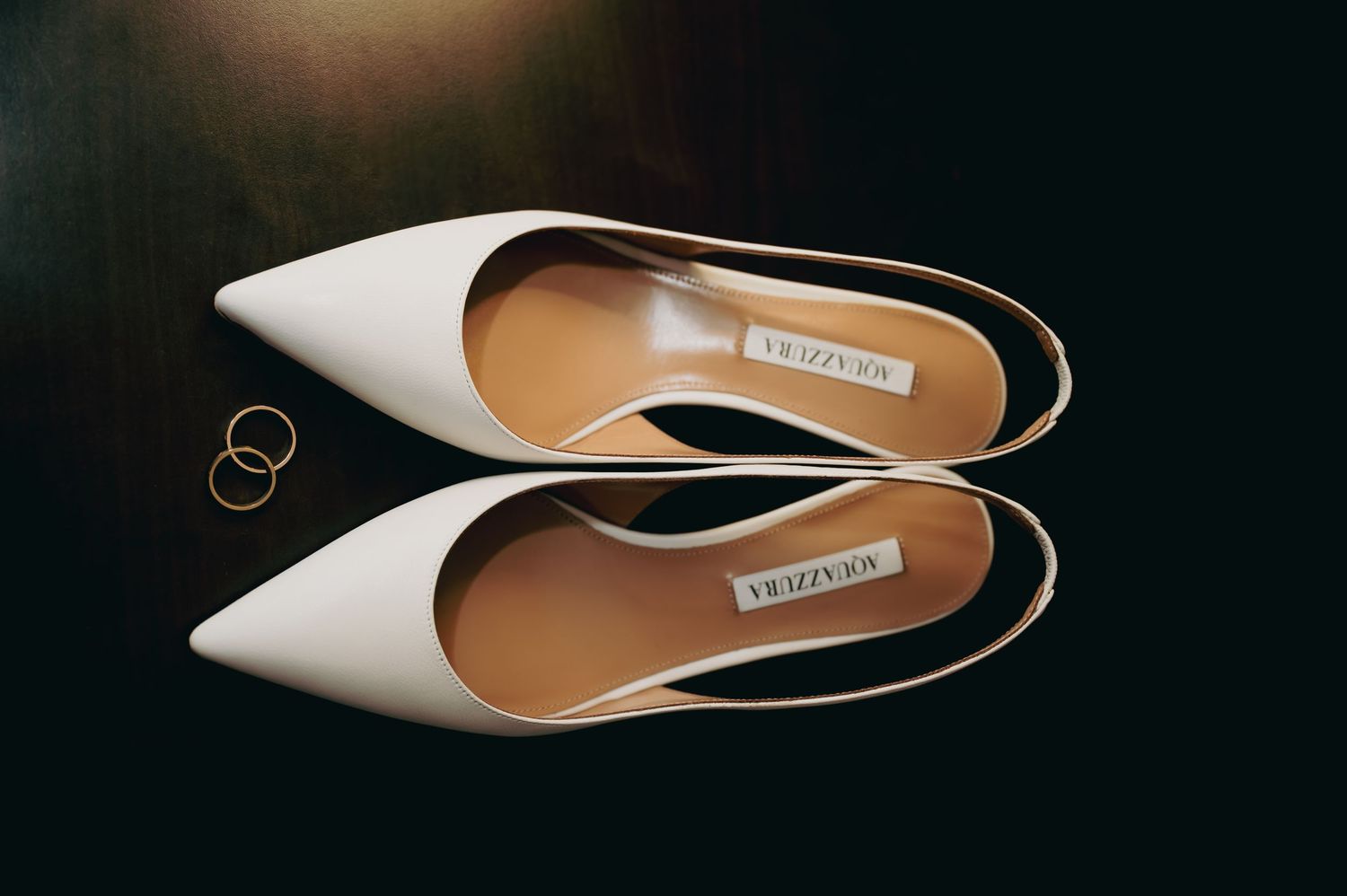White Aquazzura pointed-toe slingback flats displayed top-down with two gold wedding bands beside them.