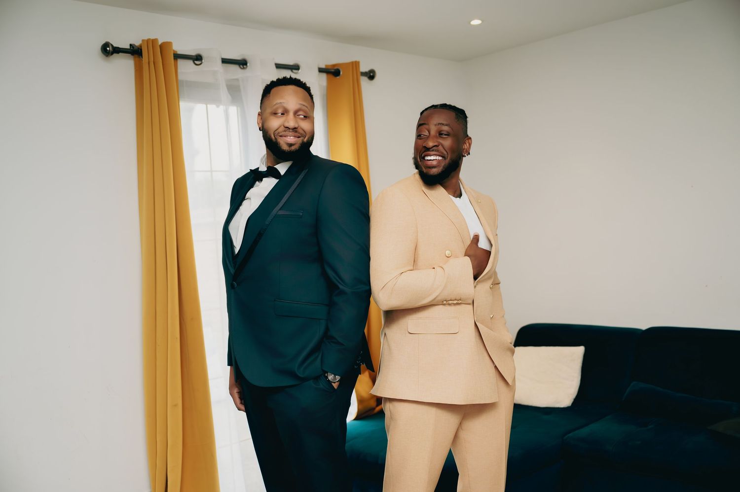 Two well-dressed grooms smile in a bright room with gold curtains, one in a teal tuxedo and one in a tan suit.