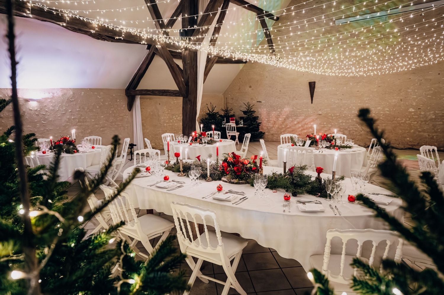 Christmas wedding banquet hall with long white tables, greenery centerpieces, red candles, and twinkling fairy lights above.