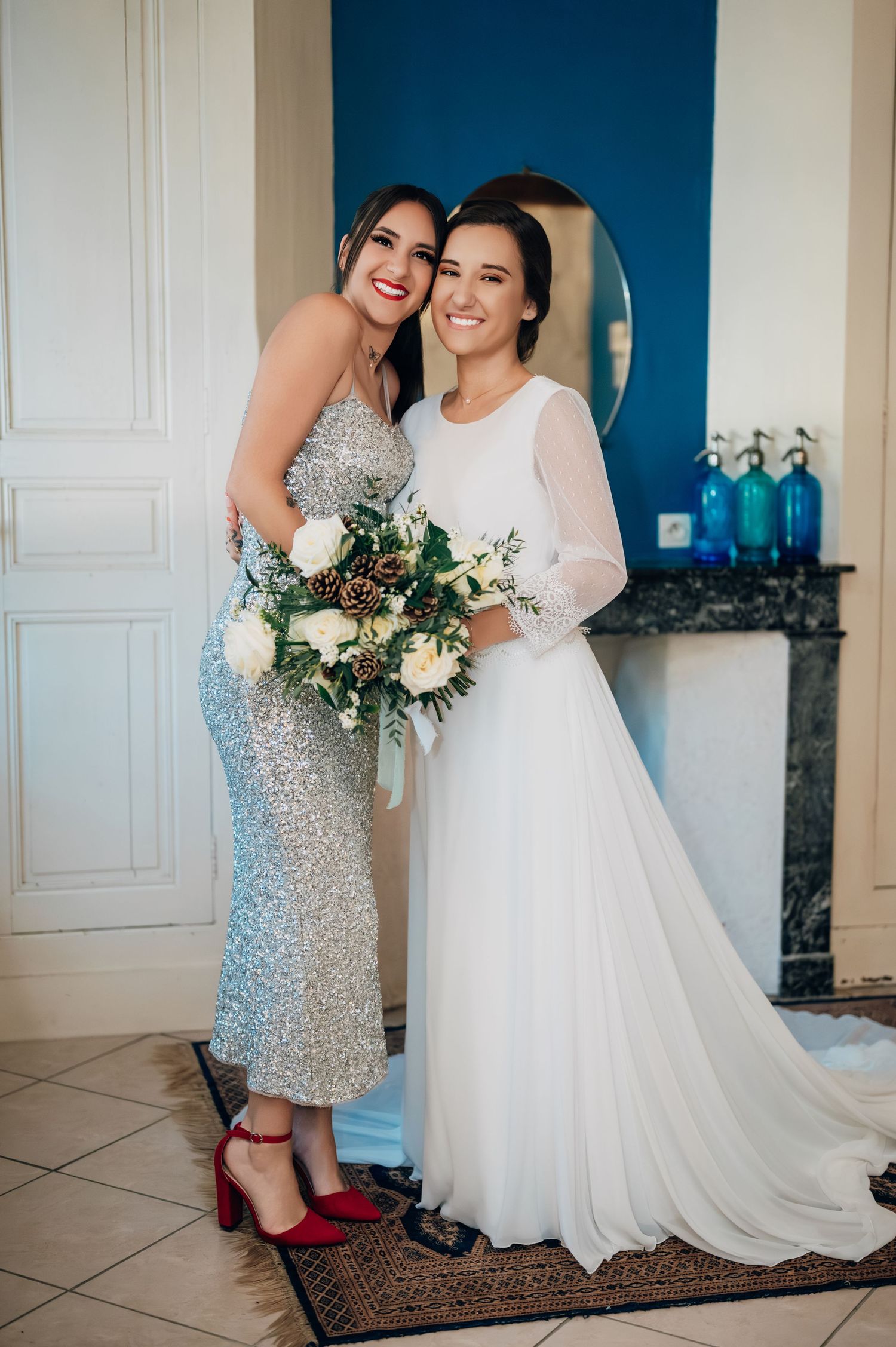 Bride in white gown smiling with bridesmaid in silver sequin dress holding winter bouquet in elegant room.