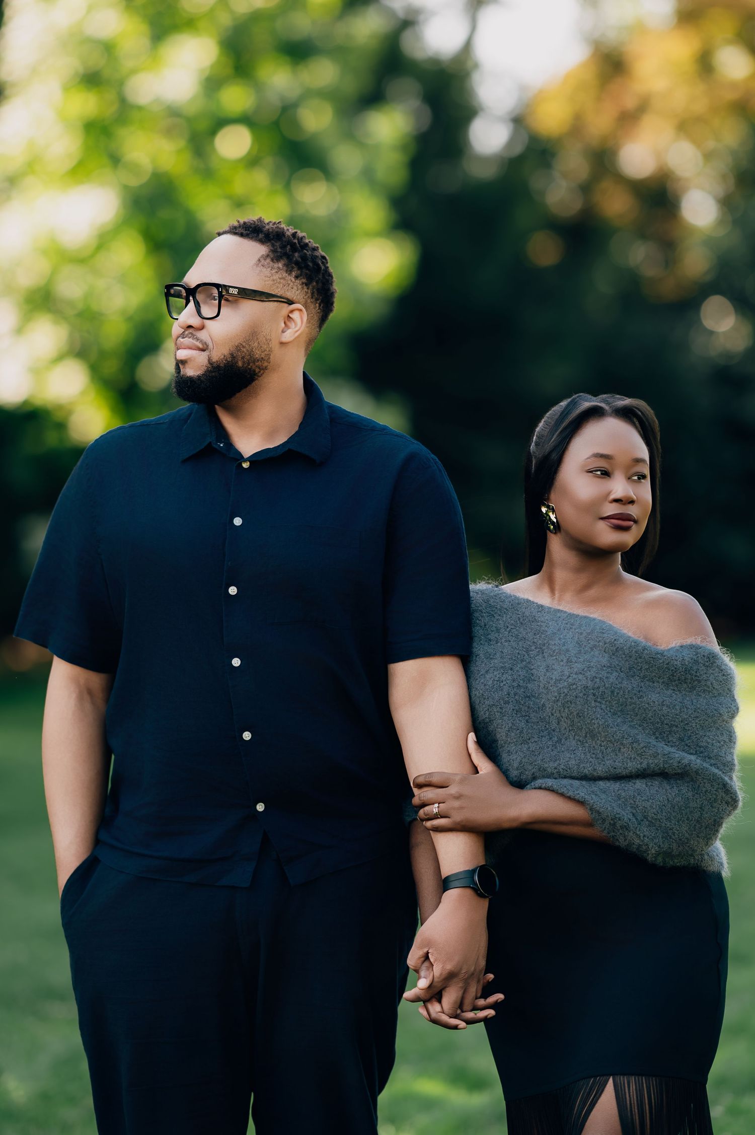 Stylish couple holds hands in a lush park, man in navy shirt, woman in gray wrap.
