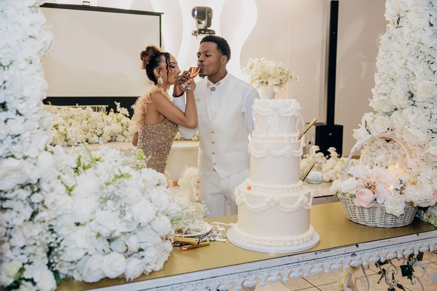 Couple shares wedding cake at elegant all-white reception surrounded by white floral arrangements and candles.