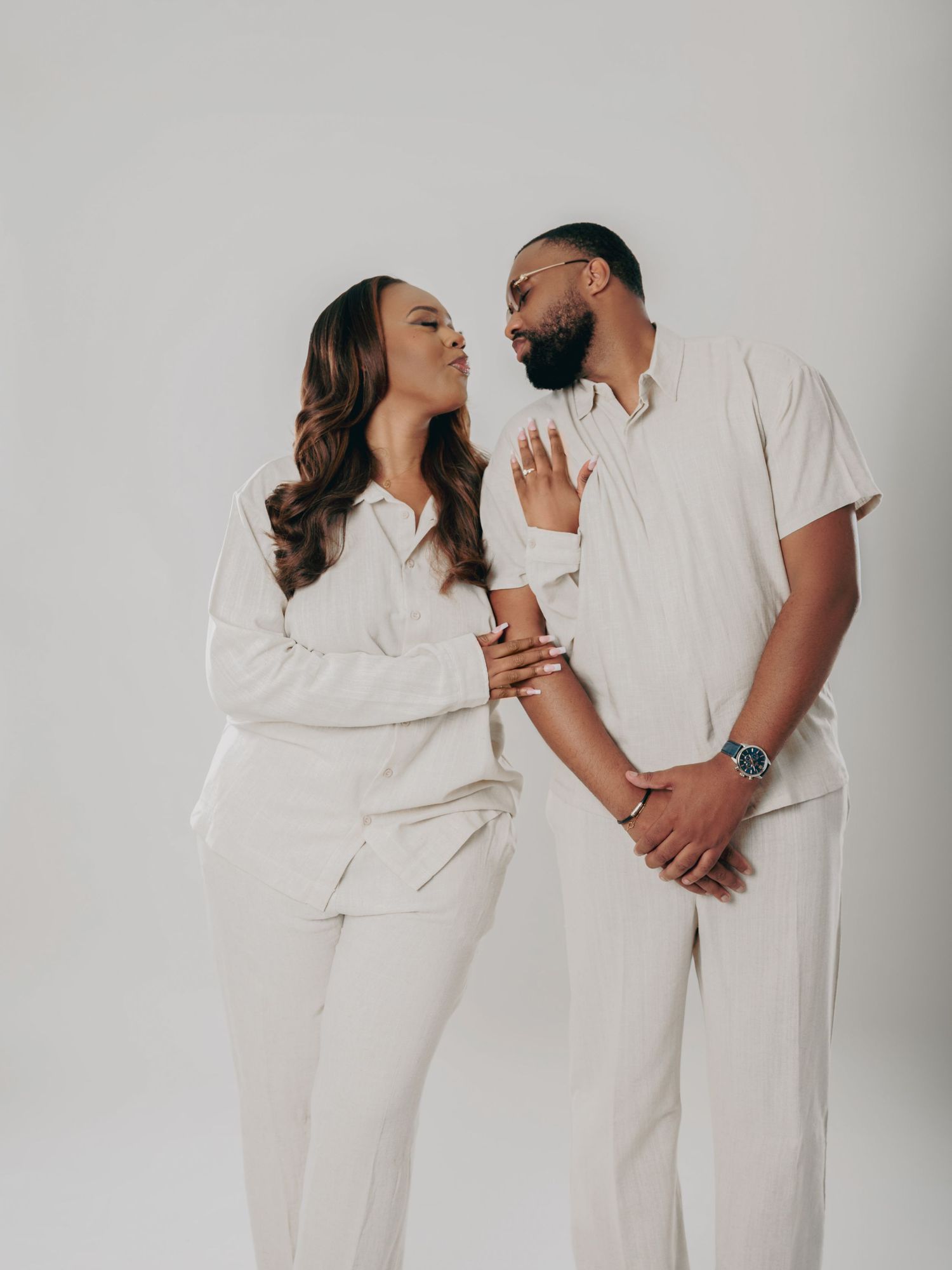 Couple in matching white outfits share a loving gaze while holding hands in a studio portrait.