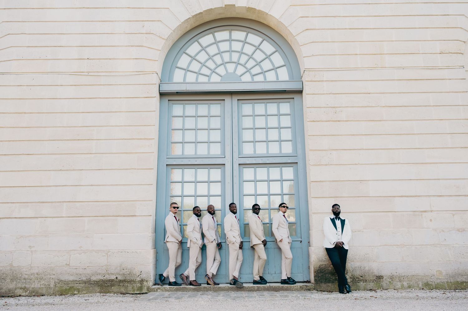 Wedding party with groomsmen in cream suits lined up against an elegant stone building.