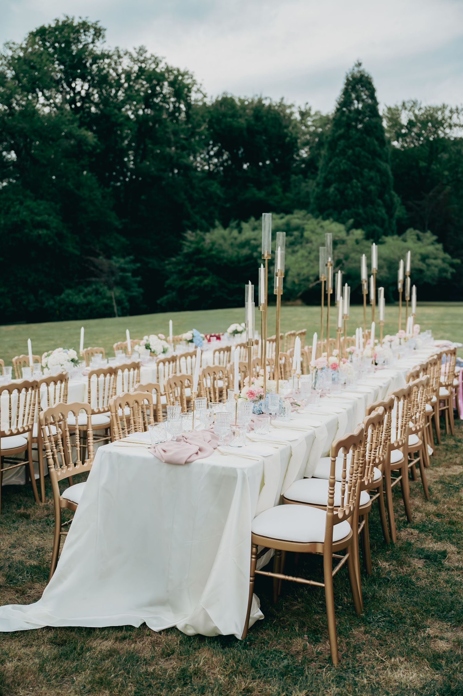 Elegant outdoor wedding reception with long white tablecloth, gold Chiavari chairs, and tall candelabras.