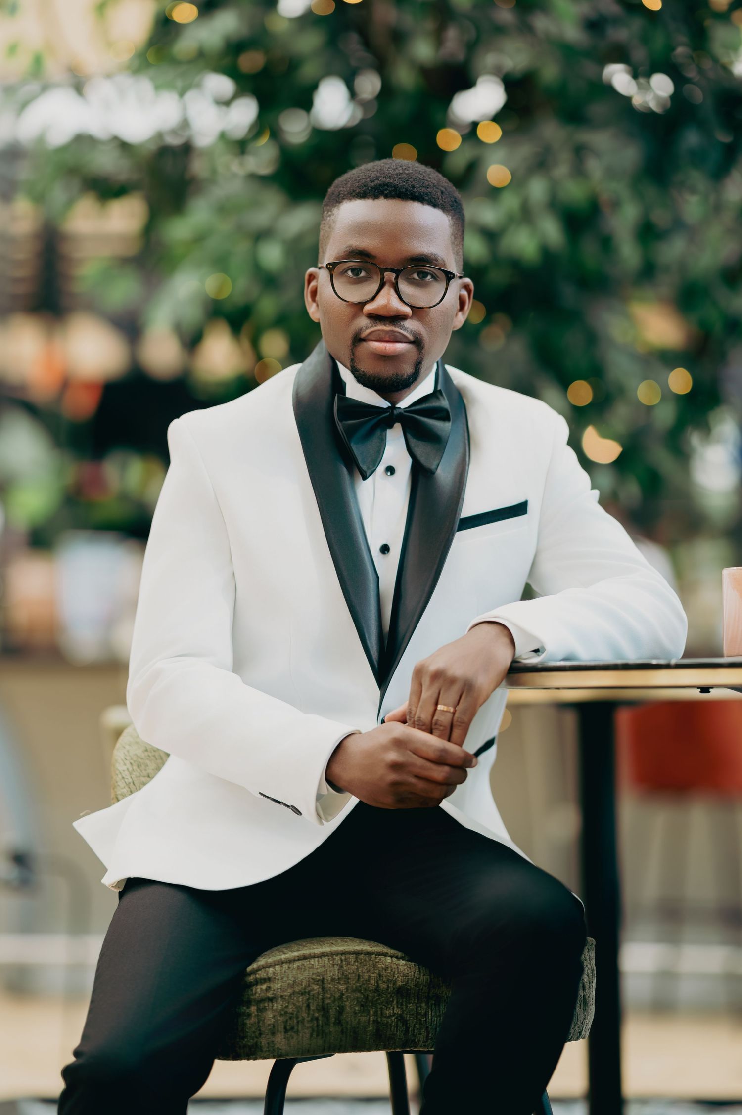 Groom in white tuxedo jacket seated at a table in a lush green outdoor setting.