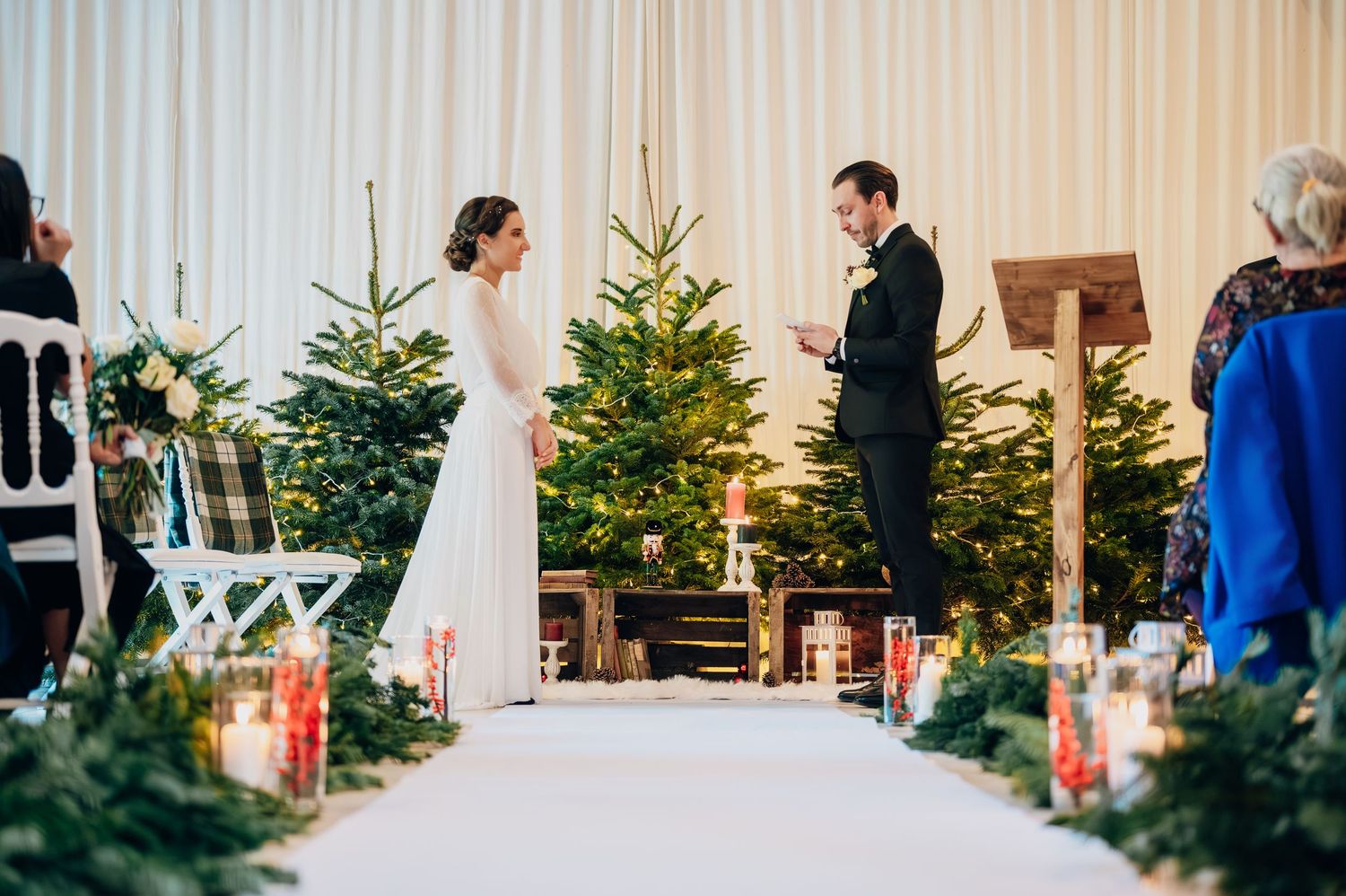 Groom reading wedding vows to bride at altar surrounded by Christmas trees and candles in barn.