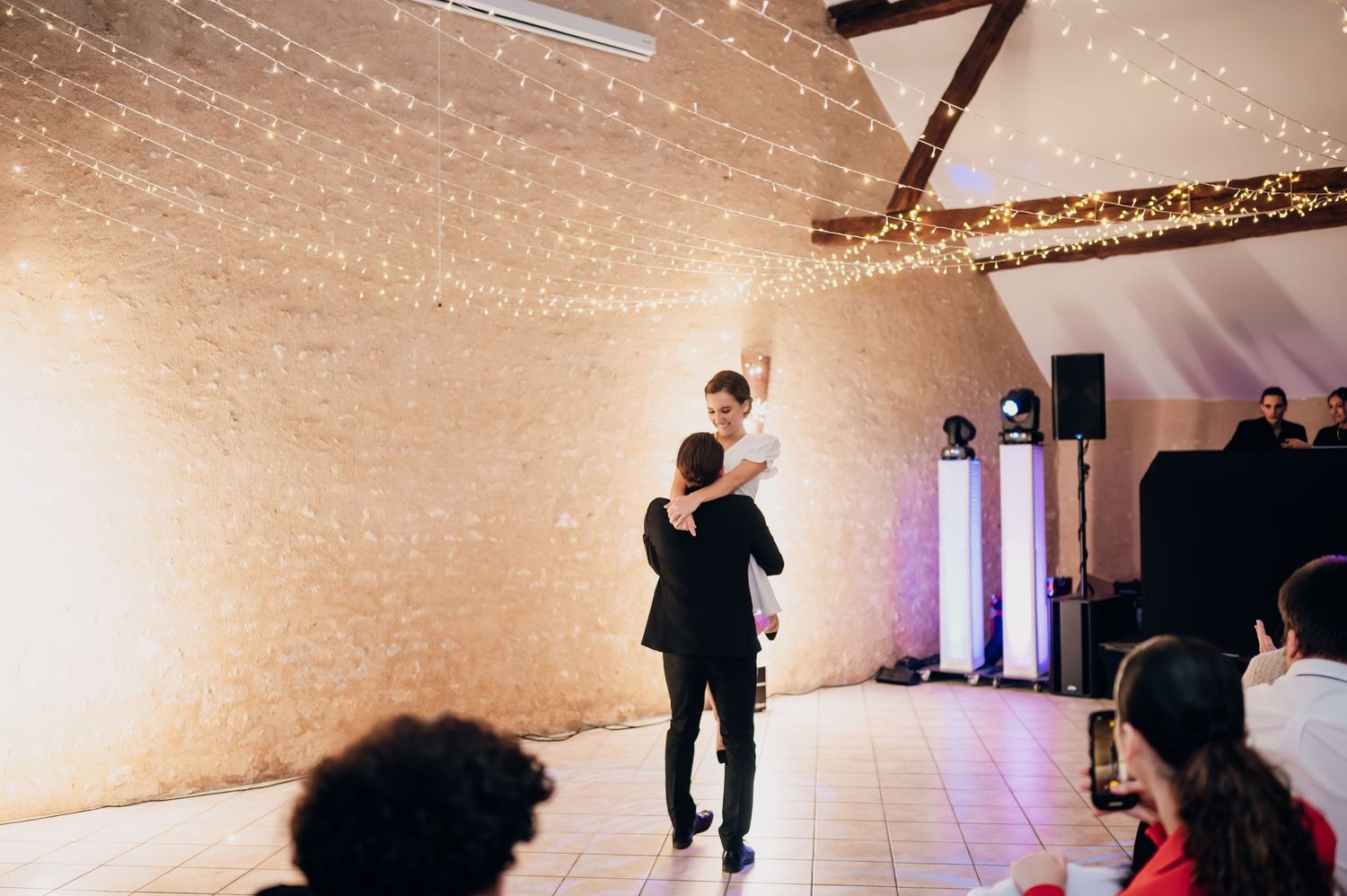 Groom lifts bride during first dance under a sparkling fairy light backdrop.