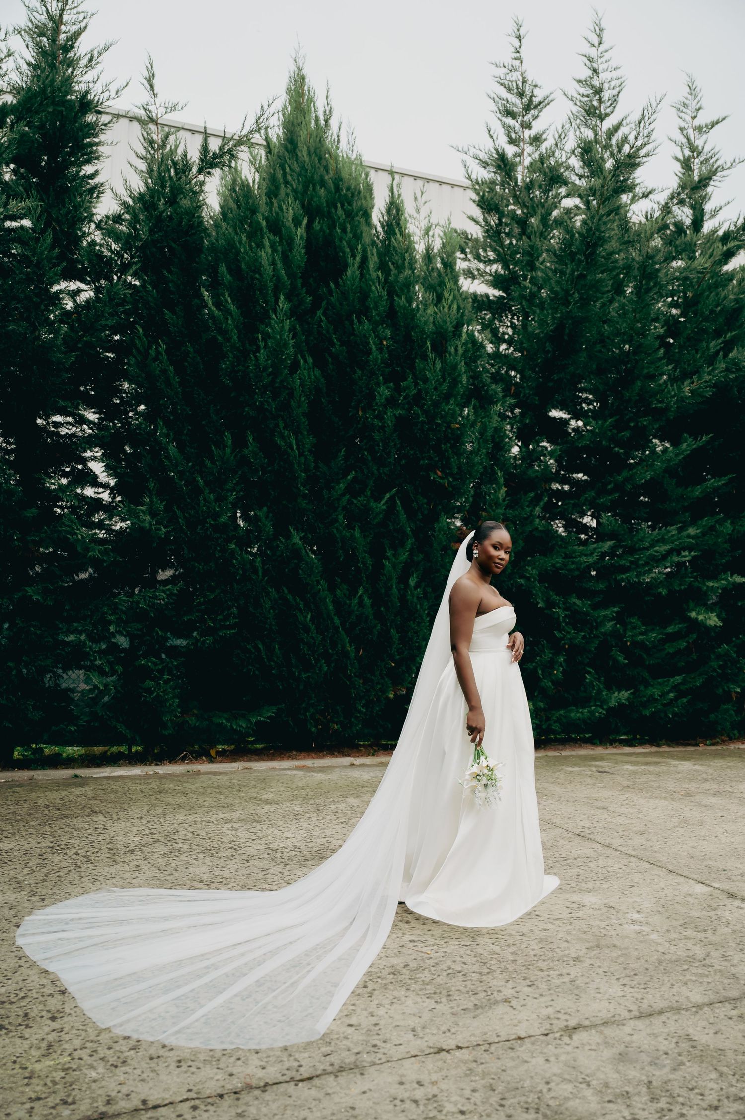 Black bride in strapless white wedding gown with dramatic cathedral veil flowing behind her near cypress trees.