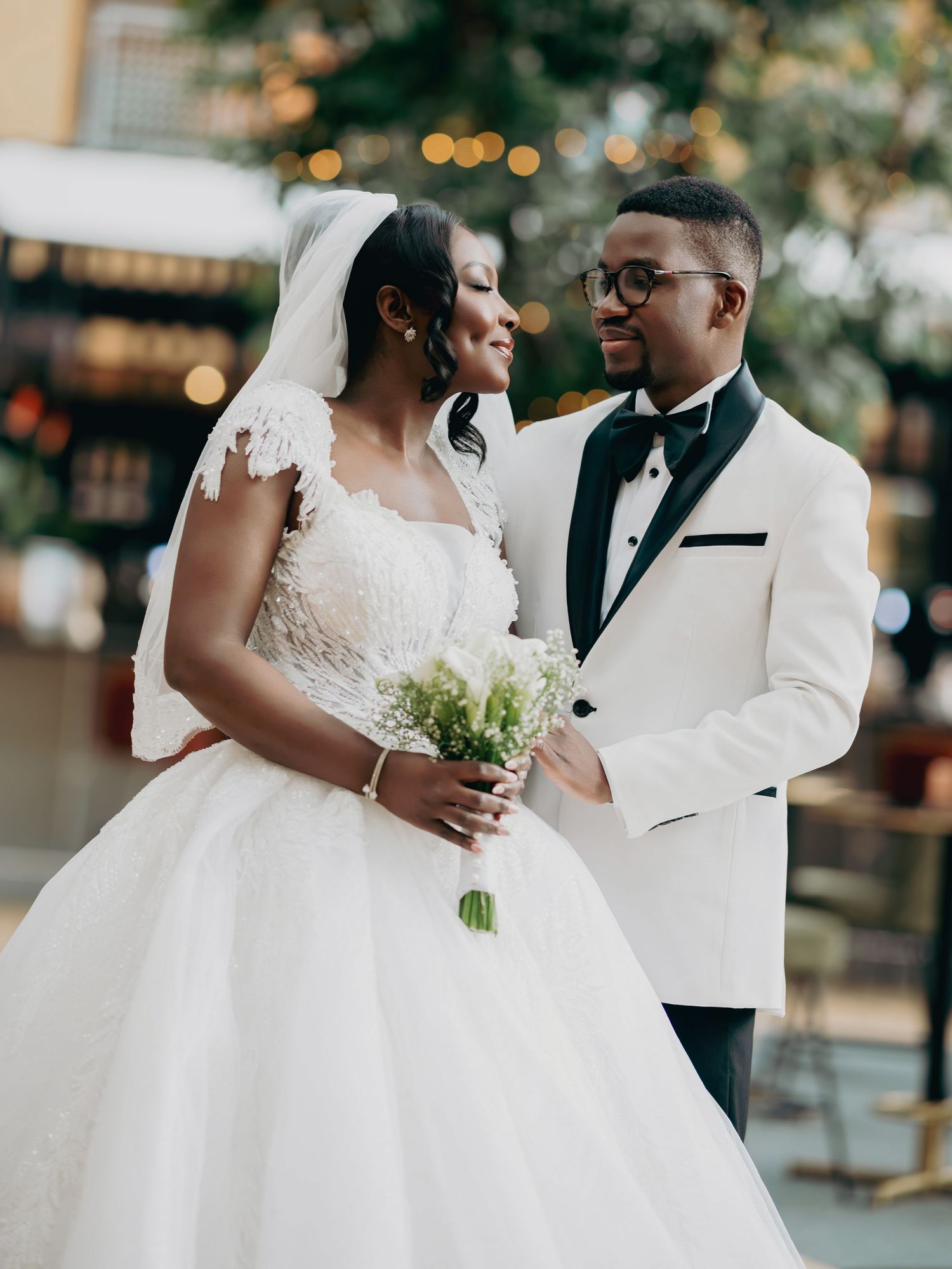 Black bride in lace ball gown and groom in white tuxedo share a loving glance at their wedding.