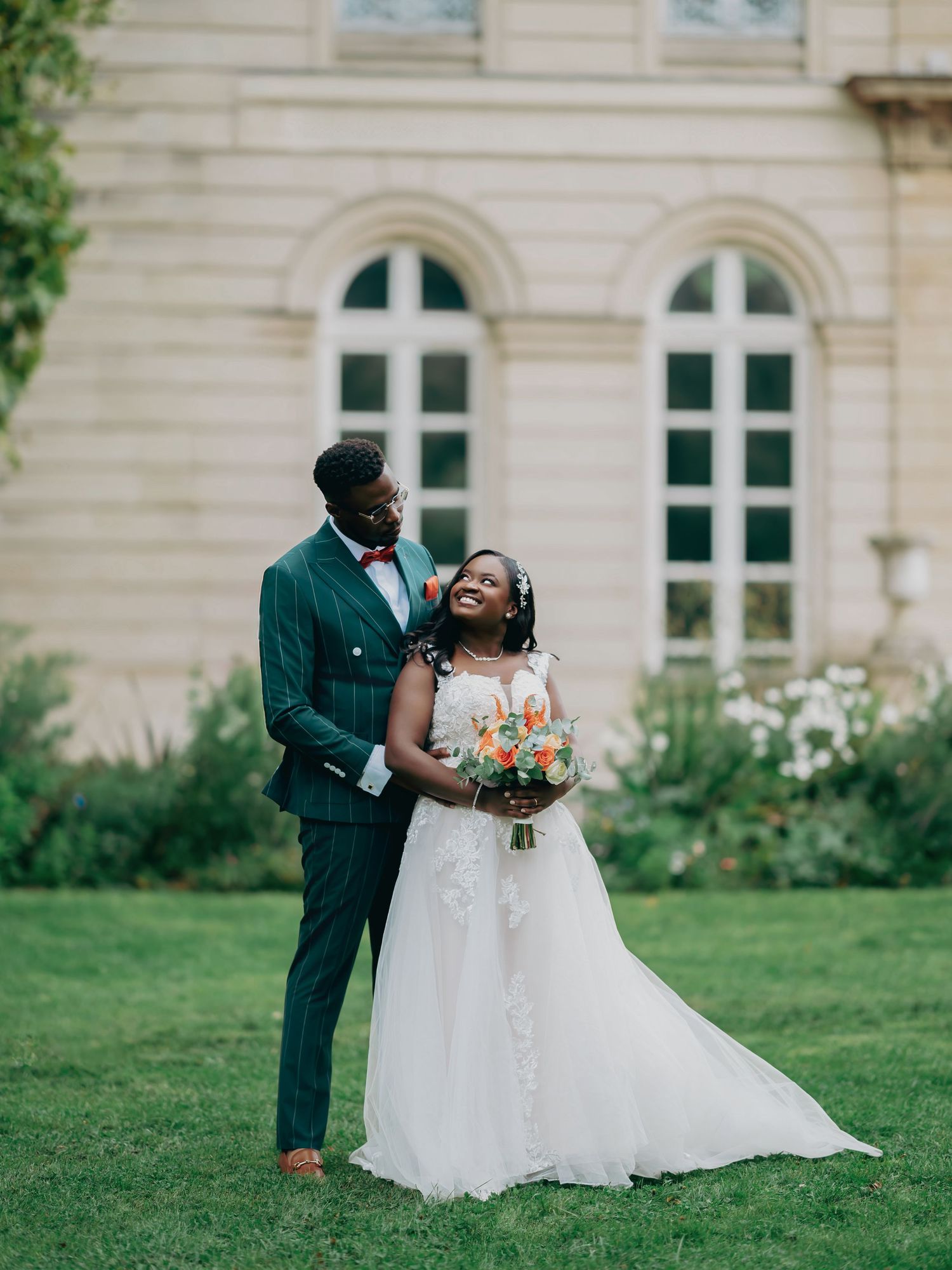Bride and groom posing together on green lawn in front of elegant stone building on wedding day.