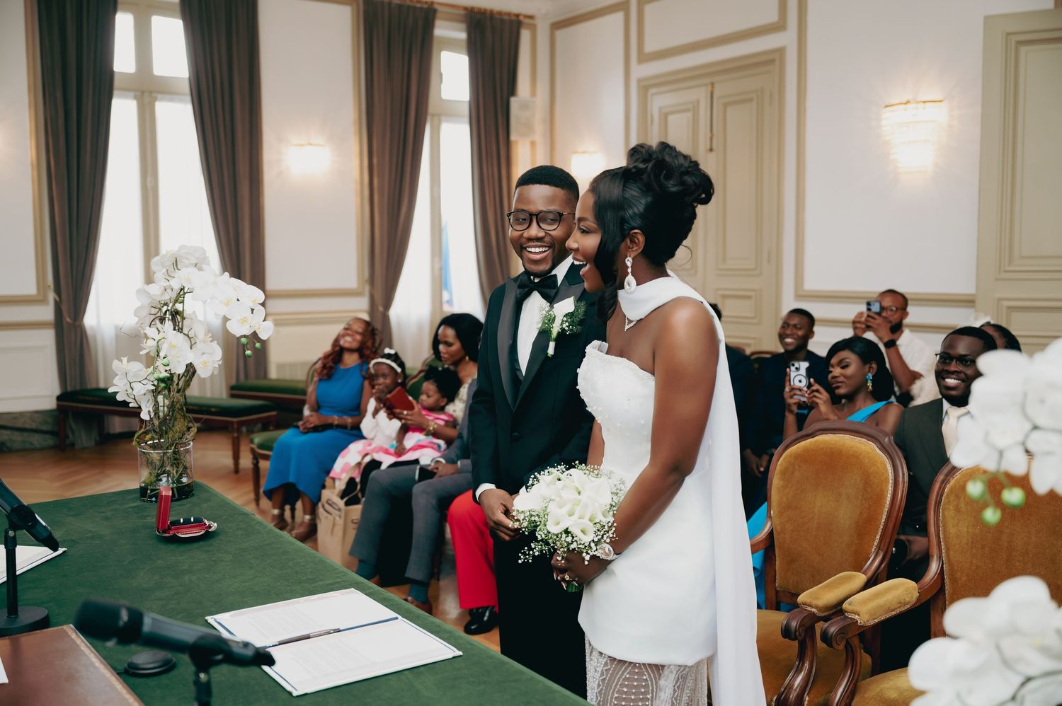Smiling bride and groom exchange vows at civil wedding ceremony with guests seated in elegant hall.