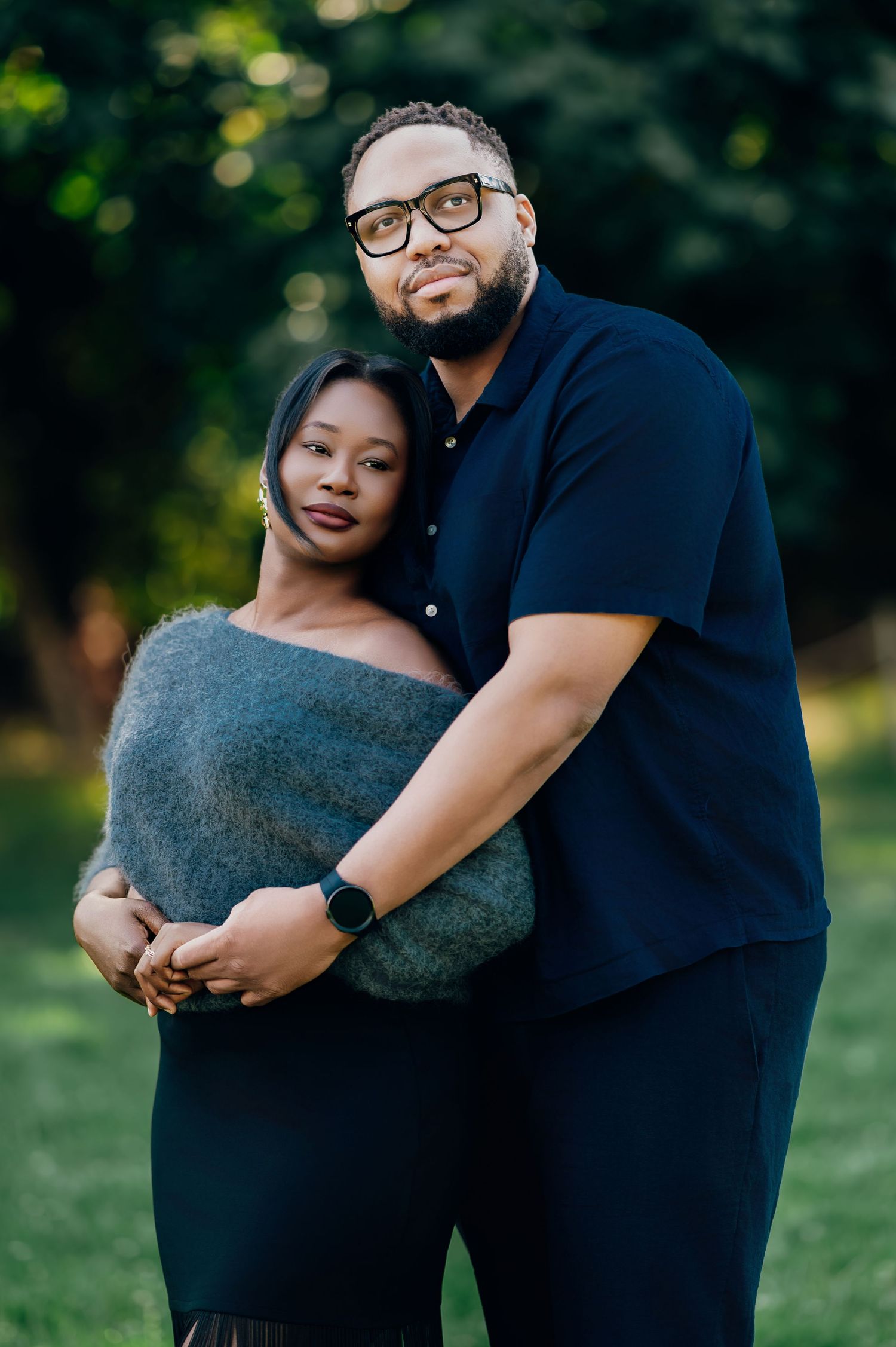 Couple poses intimately in a park, woman leaning on mans chest, both facing camera.