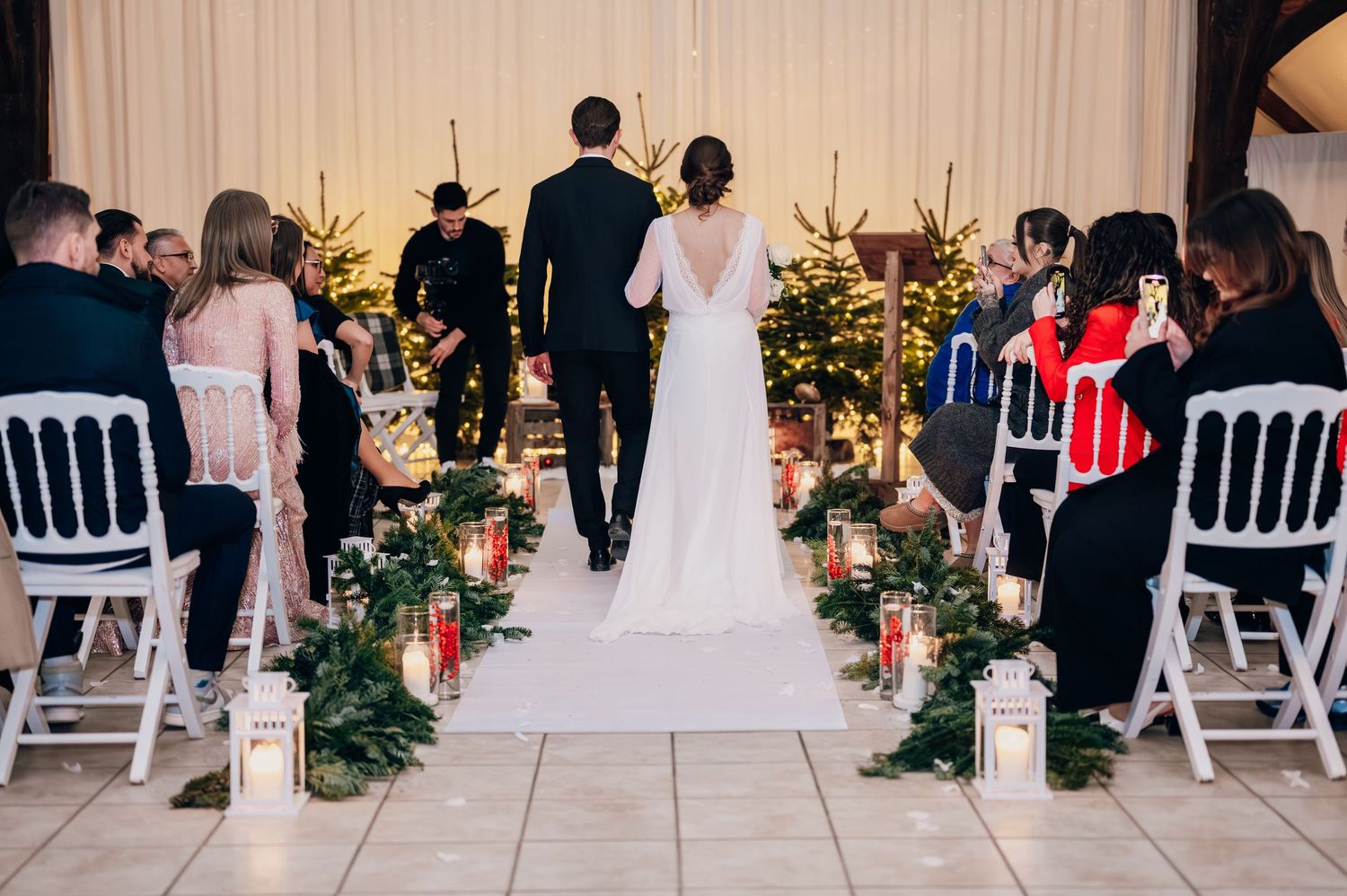 Bride and groom walking down the aisle at a Christmas wedding ceremony with greenery, lanterns, and decorated Christmas trees.