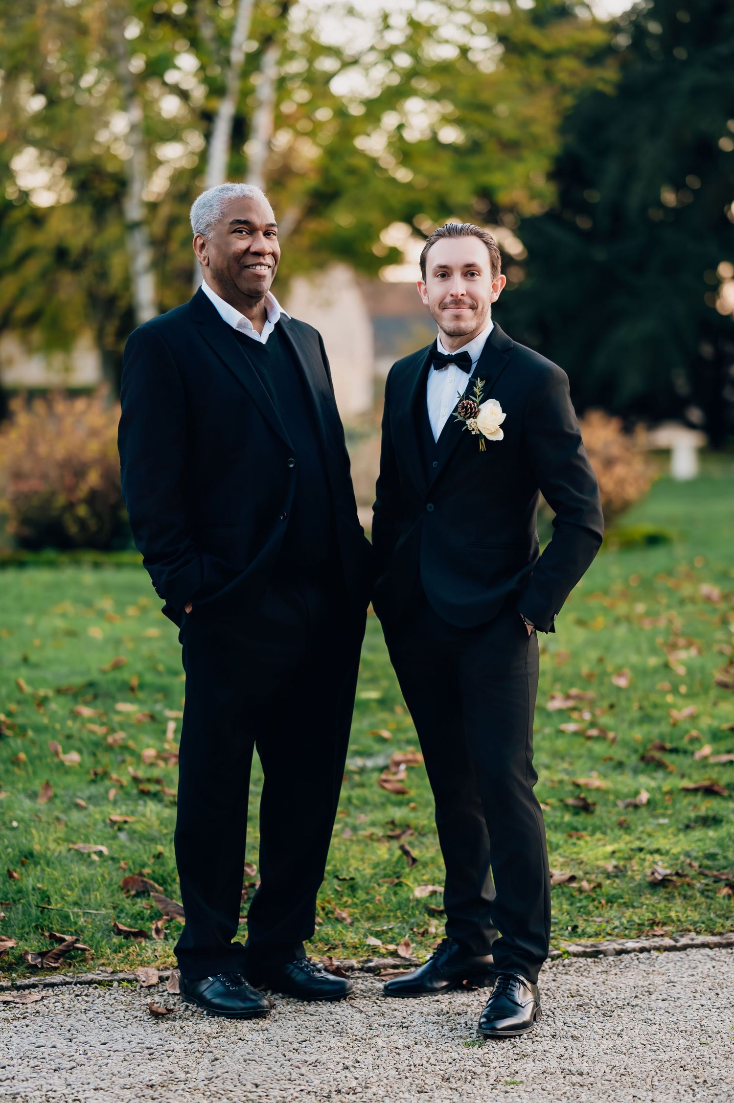 Groom in black tuxedo with boutonniere poses with older gentleman in black suit at outdoor wedding.