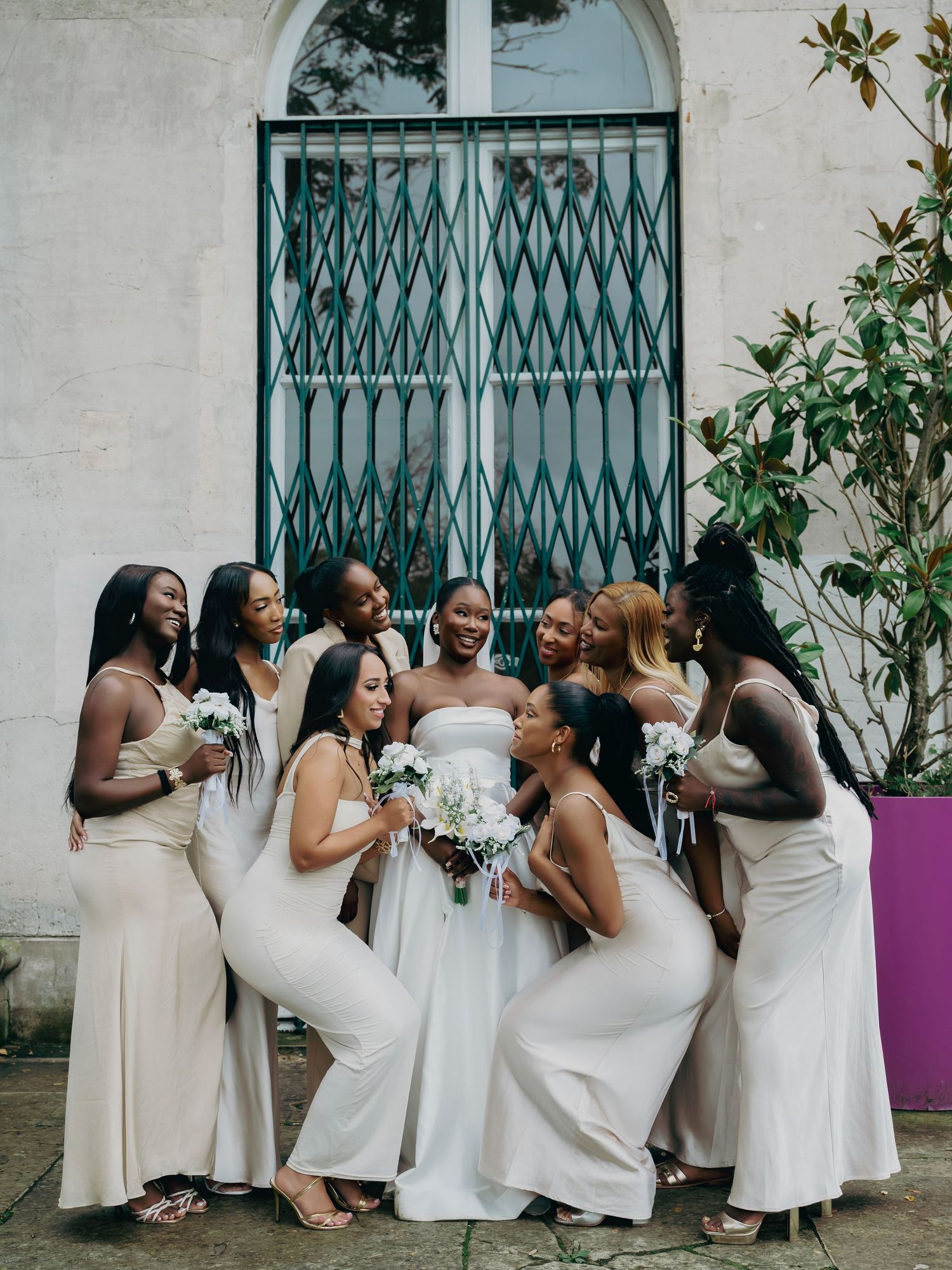 Bride in white strapless gown surrounded by bridesmaids in champagne dresses holding white bouquets outdoors.