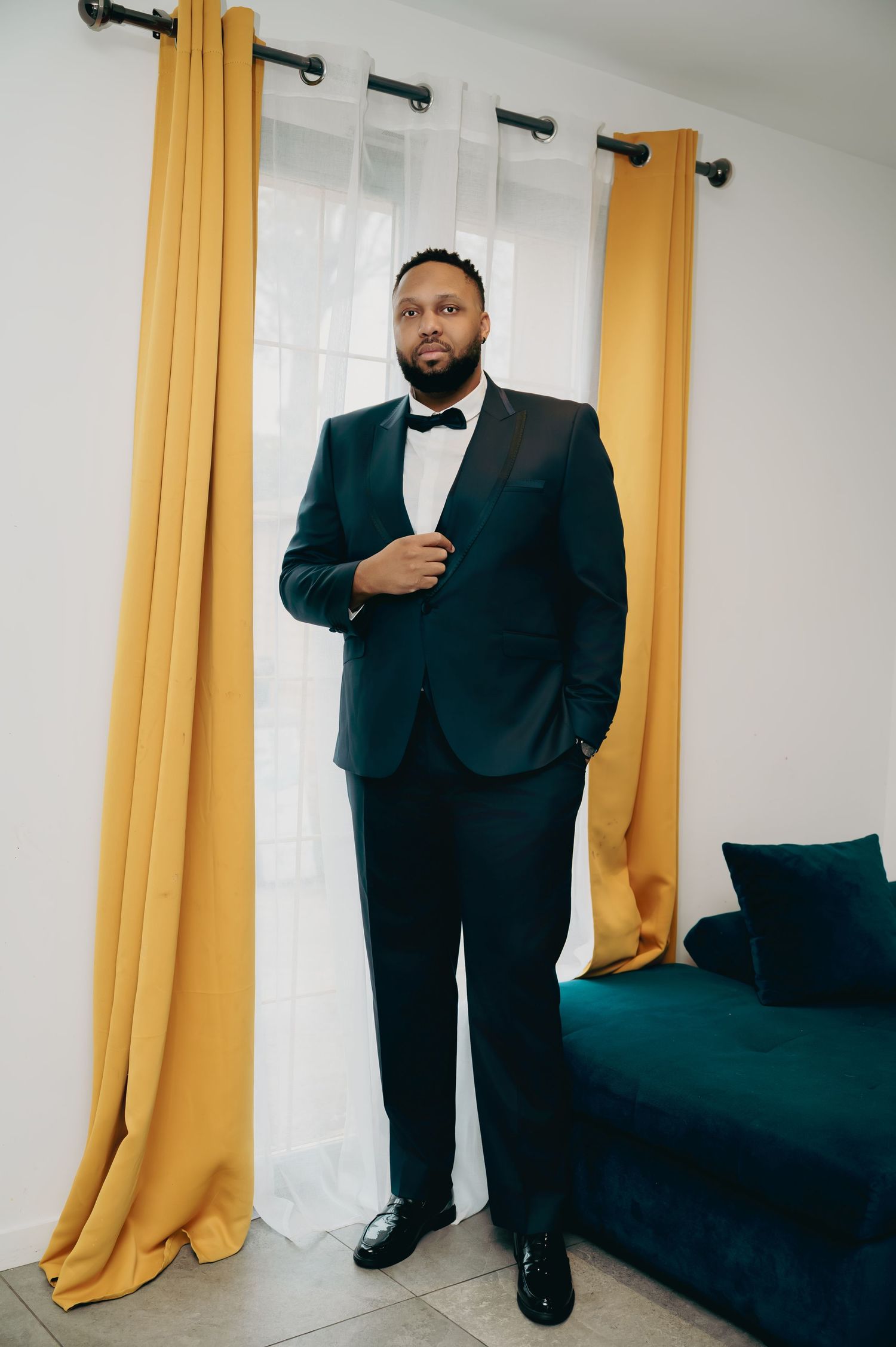 Groom in elegant dark teal tuxedo with bow tie poses confidently beside gold curtains and a velvet sofa.