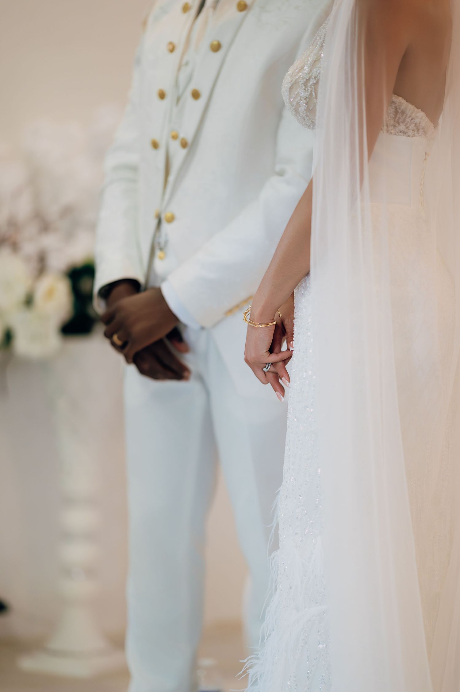 Close-up of bride and groom in white wedding attire, showcasing elegant details of lace dress and gold-buttoned suit.
