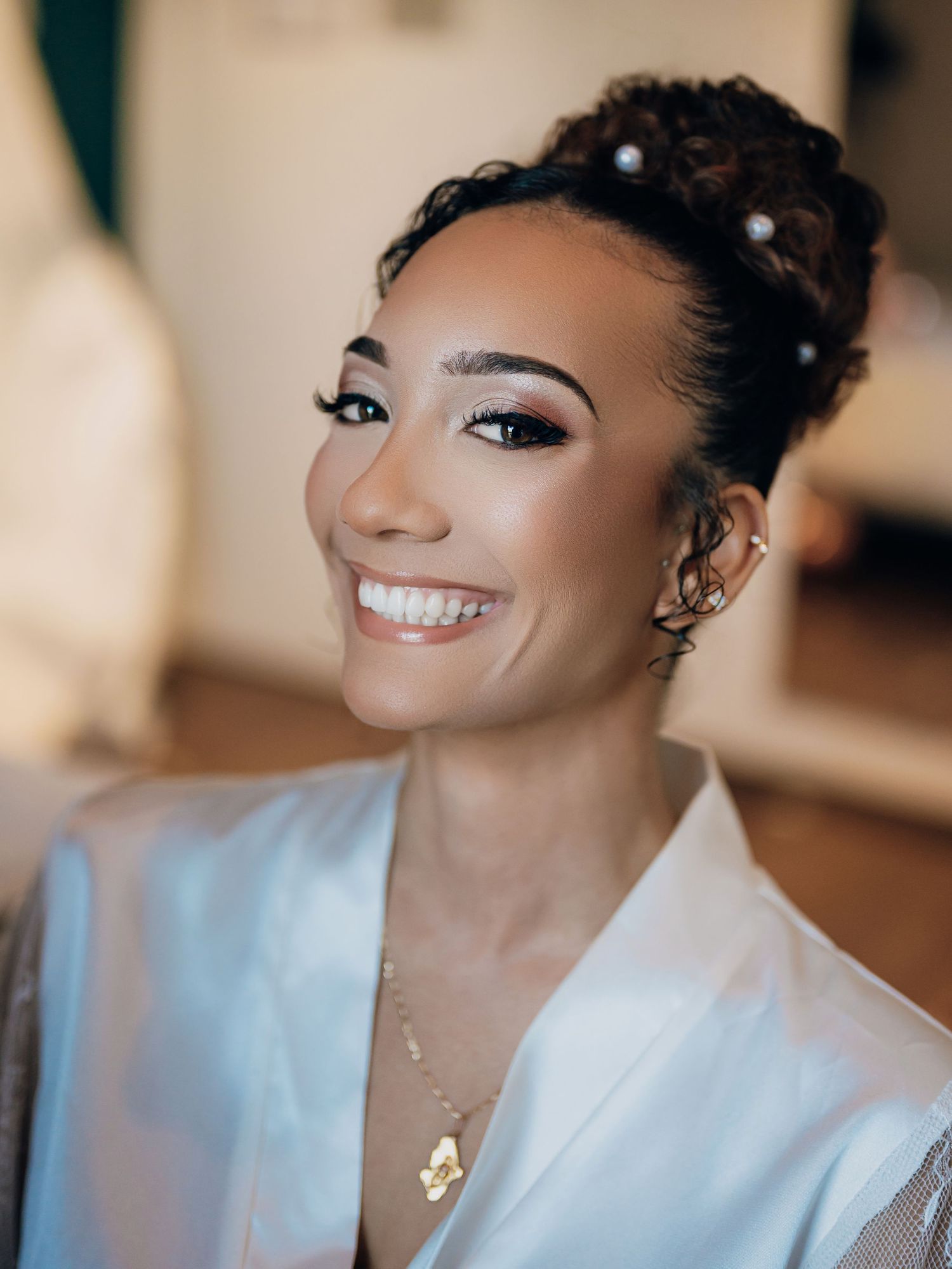 Radiant bride smiling with elegant updo, pearl hair pins, and gold necklace on wedding day.