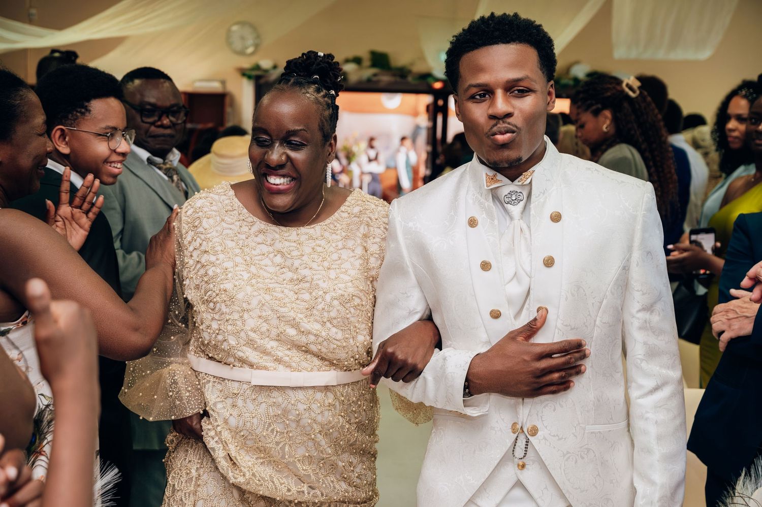 Groom in white suit walking with older woman in gold dress at wedding reception celebration.