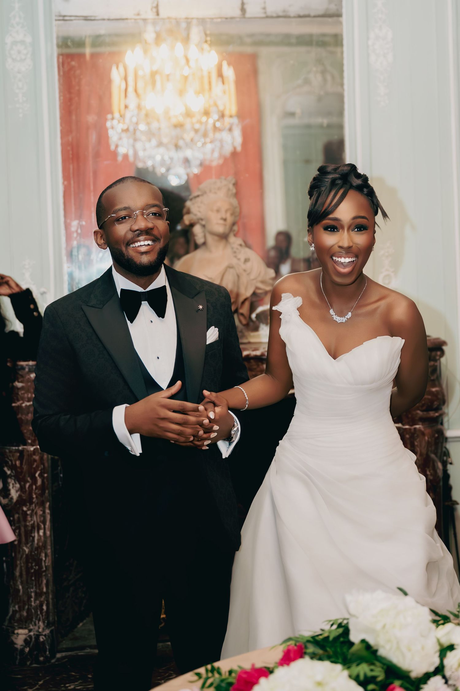 Elegant bride and groom smile while walking arm-in-arm at their wedding reception with chandelier and floral decor.