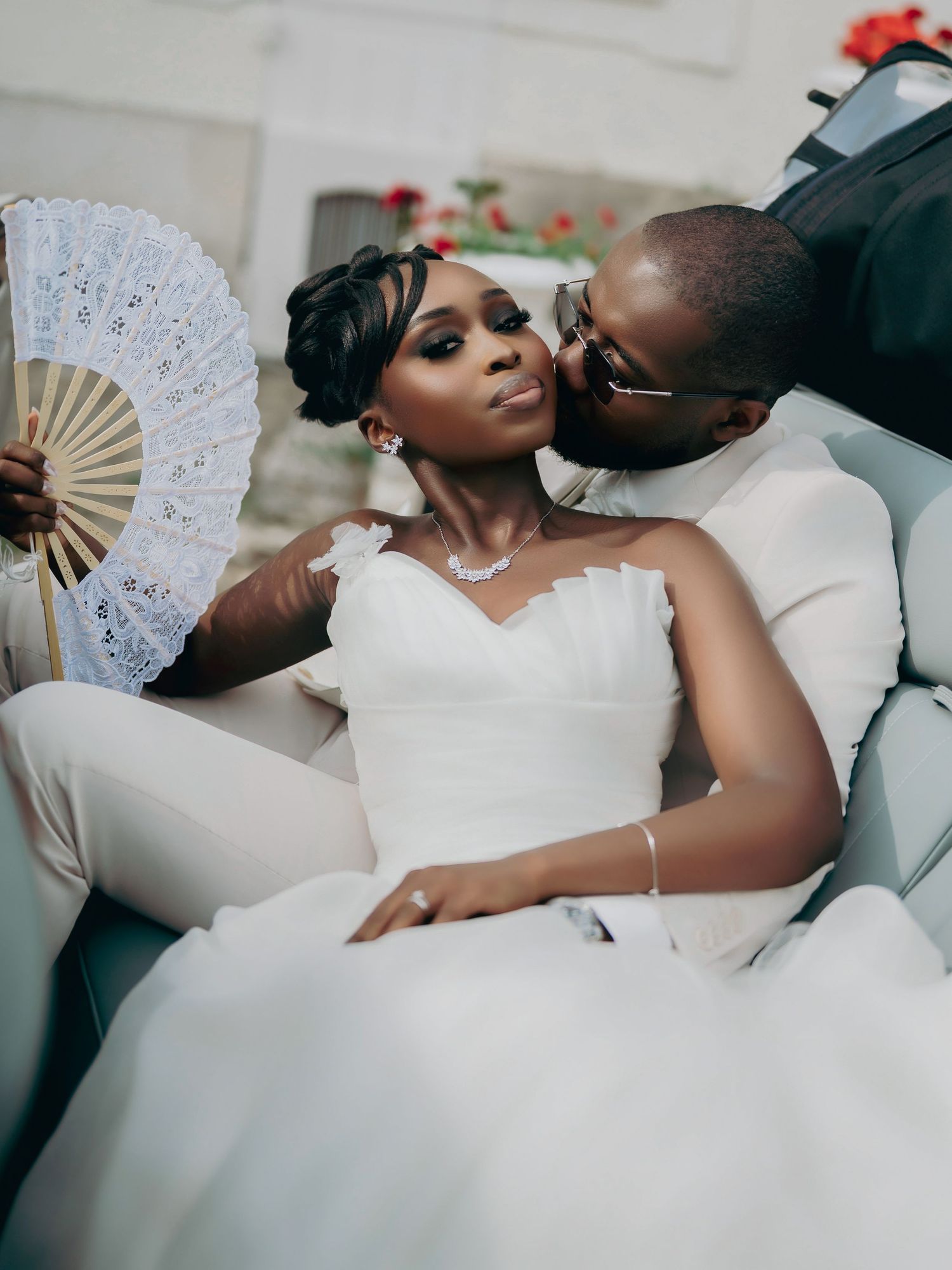 Bride holding lace fan as groom kisses her cheek while relaxing together in vintage convertible car.