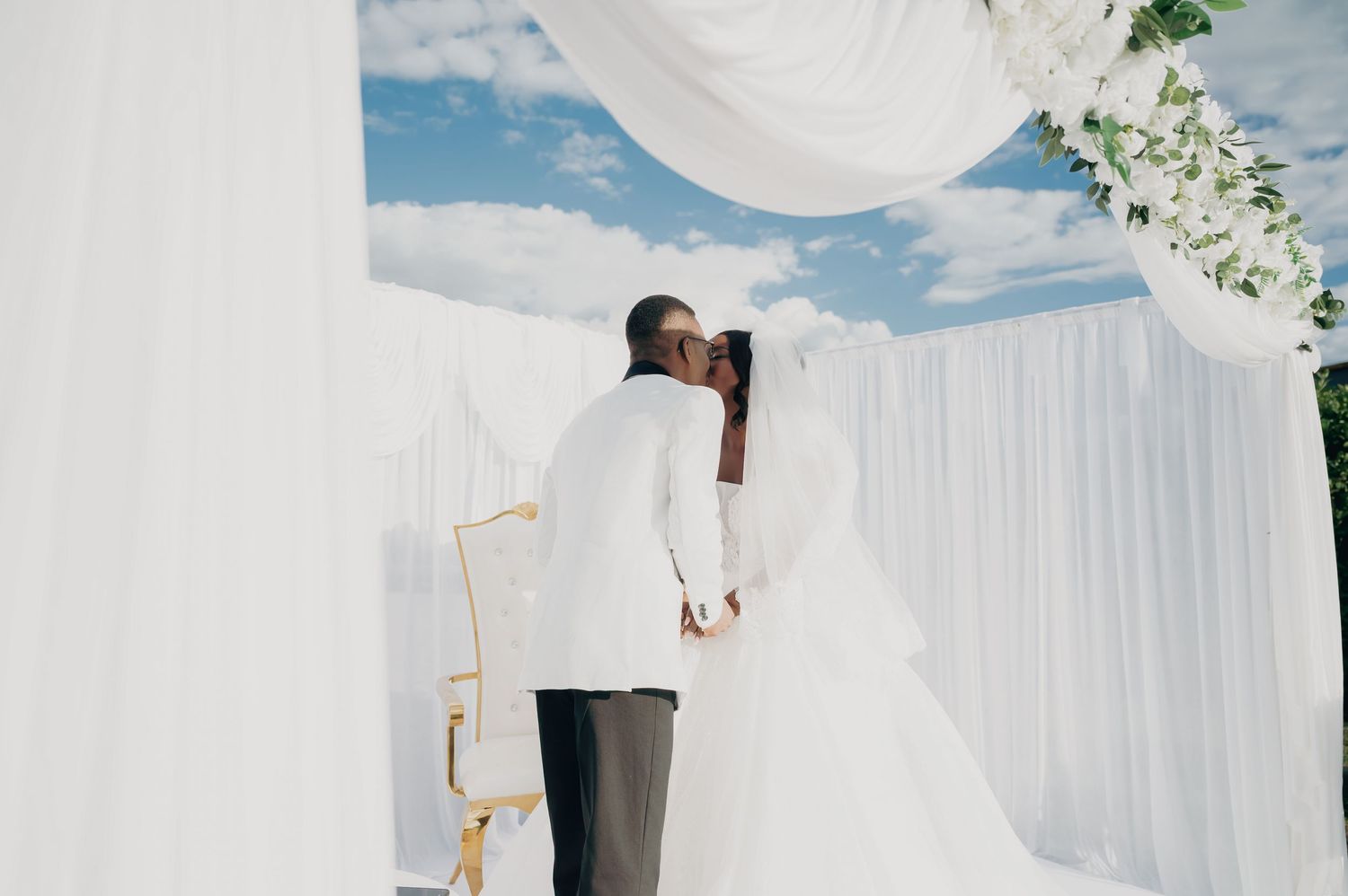 Bride and groom sharing a kiss under a white draped wedding arch with floral decorations.