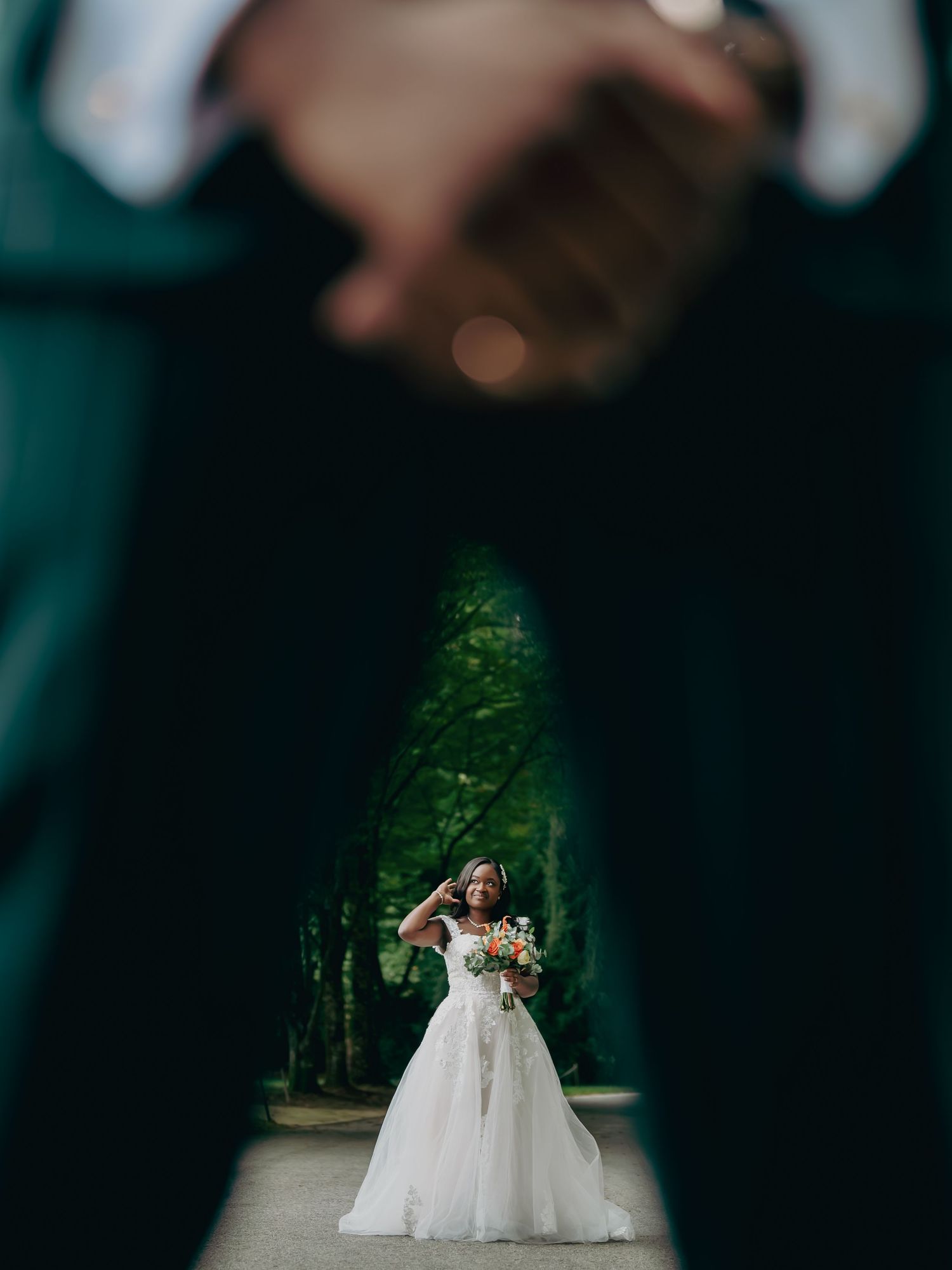 Creative wedding photo framing bride in white gown through groom's legs in lush garden setting.