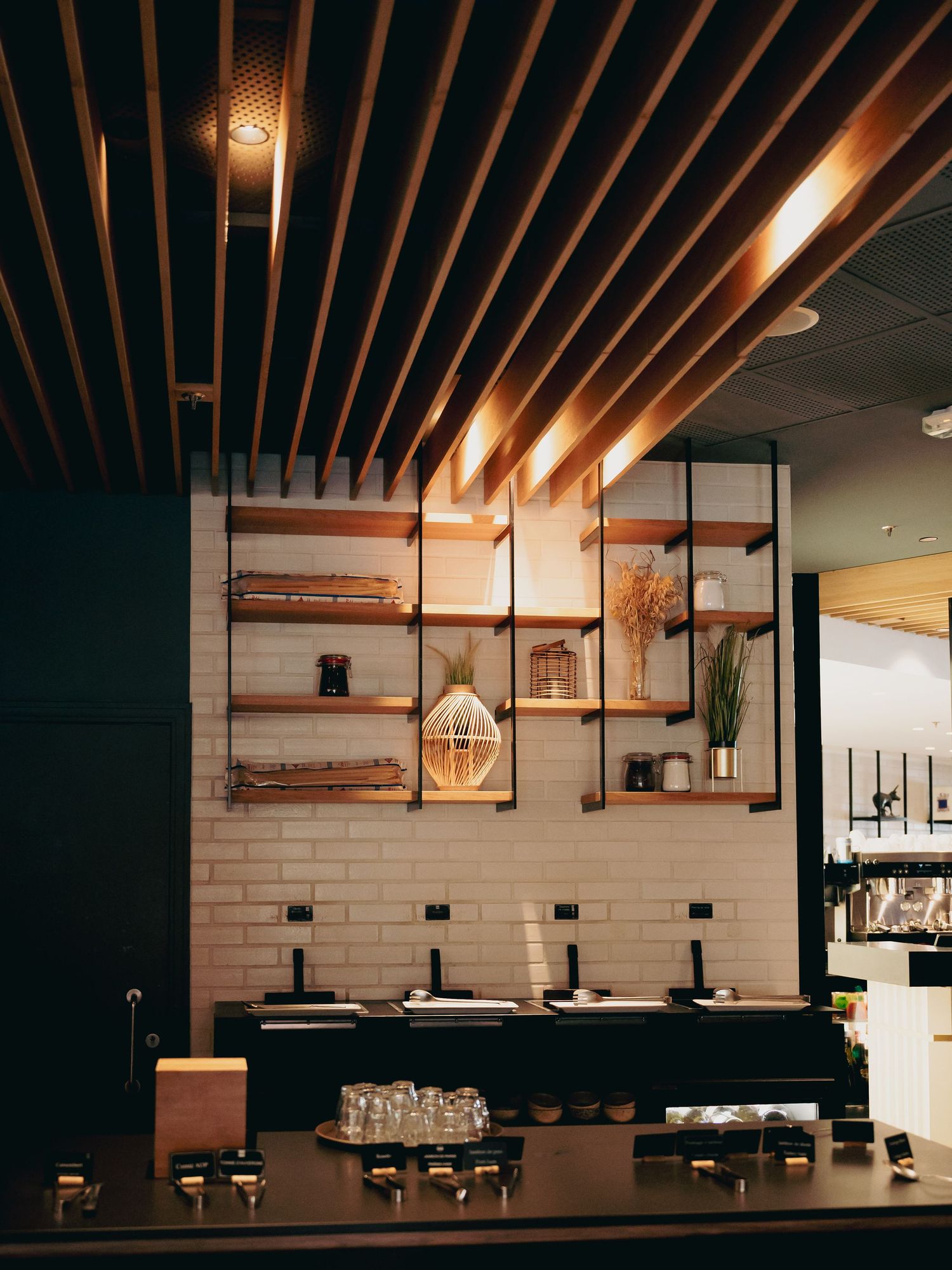 Modern restaurant bar interior with wooden slat ceiling and industrial open shelving.