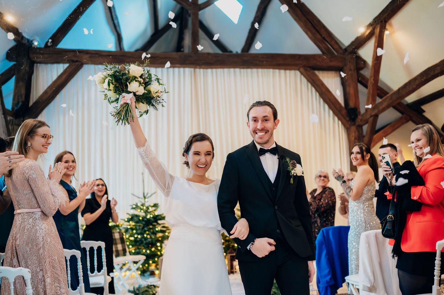 Beaming newlyweds exit ceremony as bride triumphantly raises white floral bouquet in barn.