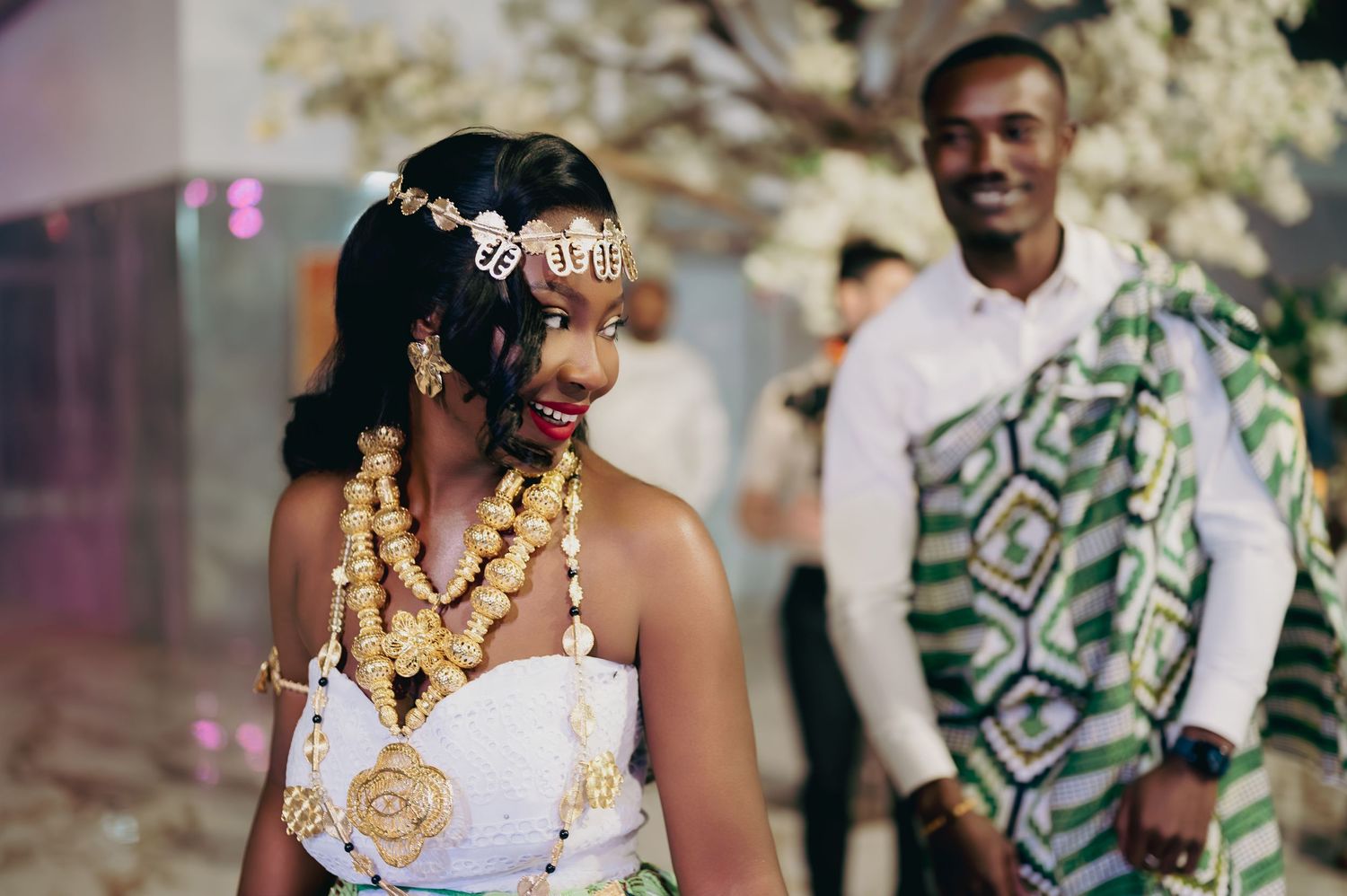 African bride adorned with gold jewelry and white traditional dress smiles at her groom during wedding ceremony.