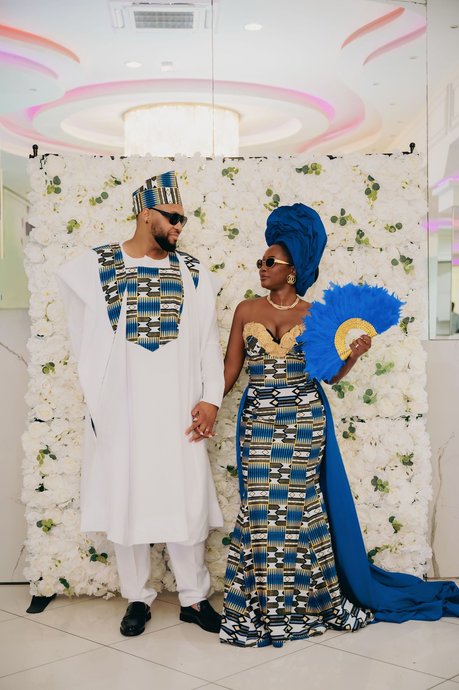 Couple in matching African kente print wedding attire pose before white floral backdrop.