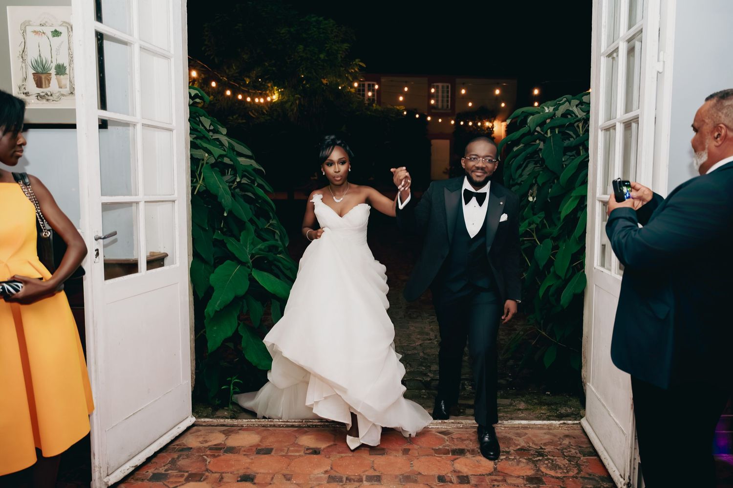 Newlywed couple makes grand entrance through French doors at outdoor wedding reception with string lights.