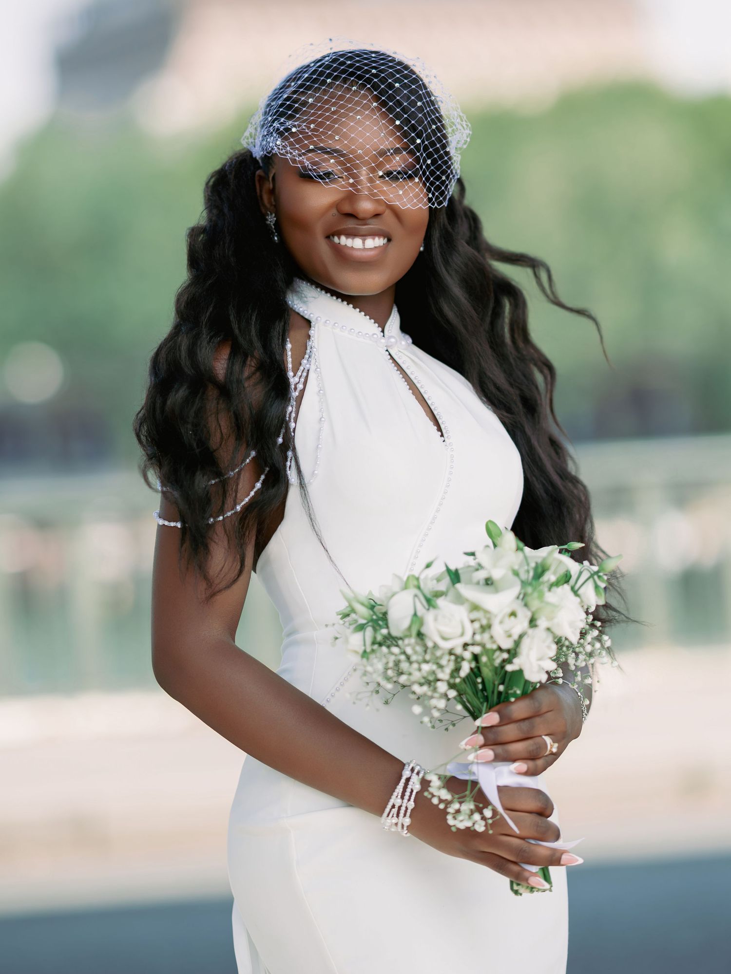 Radiant Black bride in white halter gown with birdcage veil holds white rose bouquet near the Eiffel Tower.