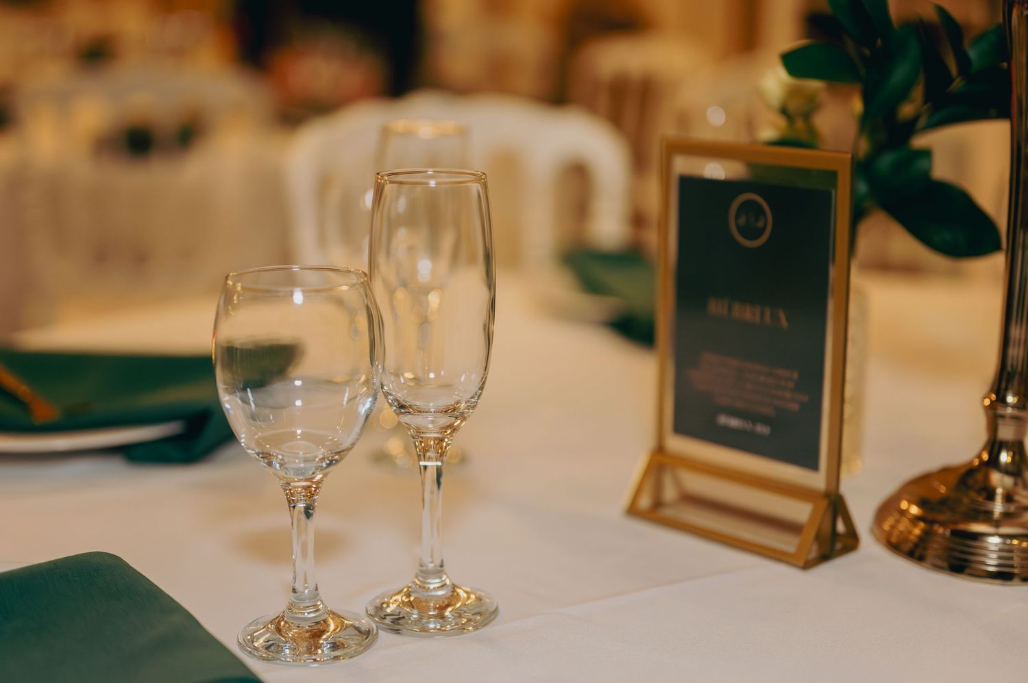 Two crystal wine glasses beside a gold-framed menu card on a white table with green napkins.