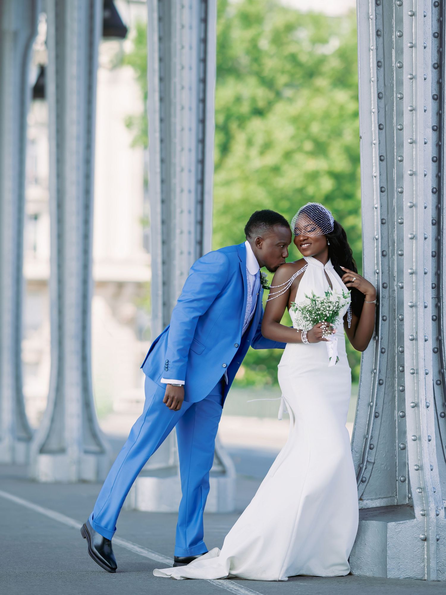 Groom in blue suit kissing bride's neck as she laughs near metal columns in outdoor wedding portrait.