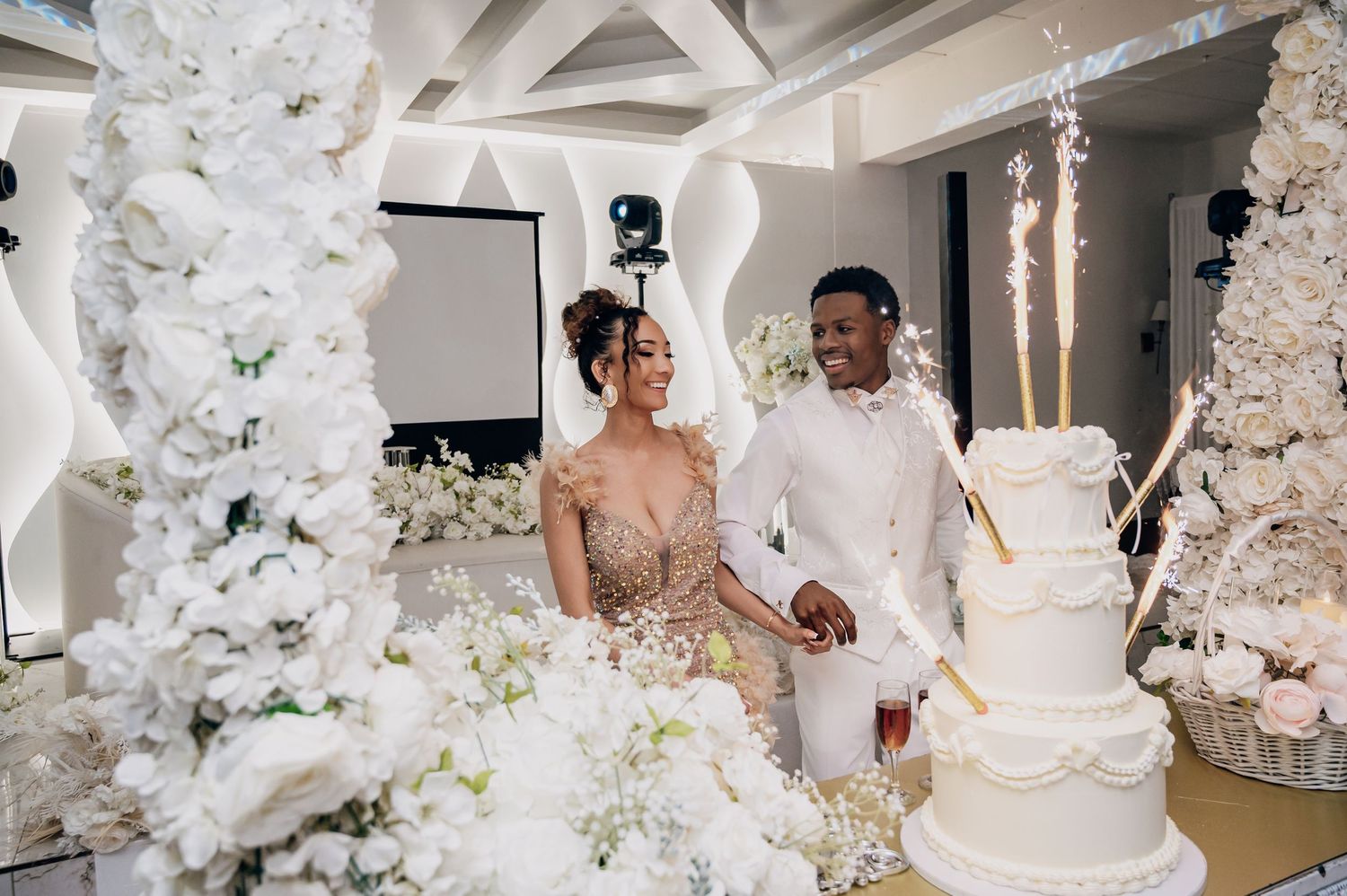 Couple cuts tiered white wedding cake surrounded by white floral arrangements with sparklers at reception.
