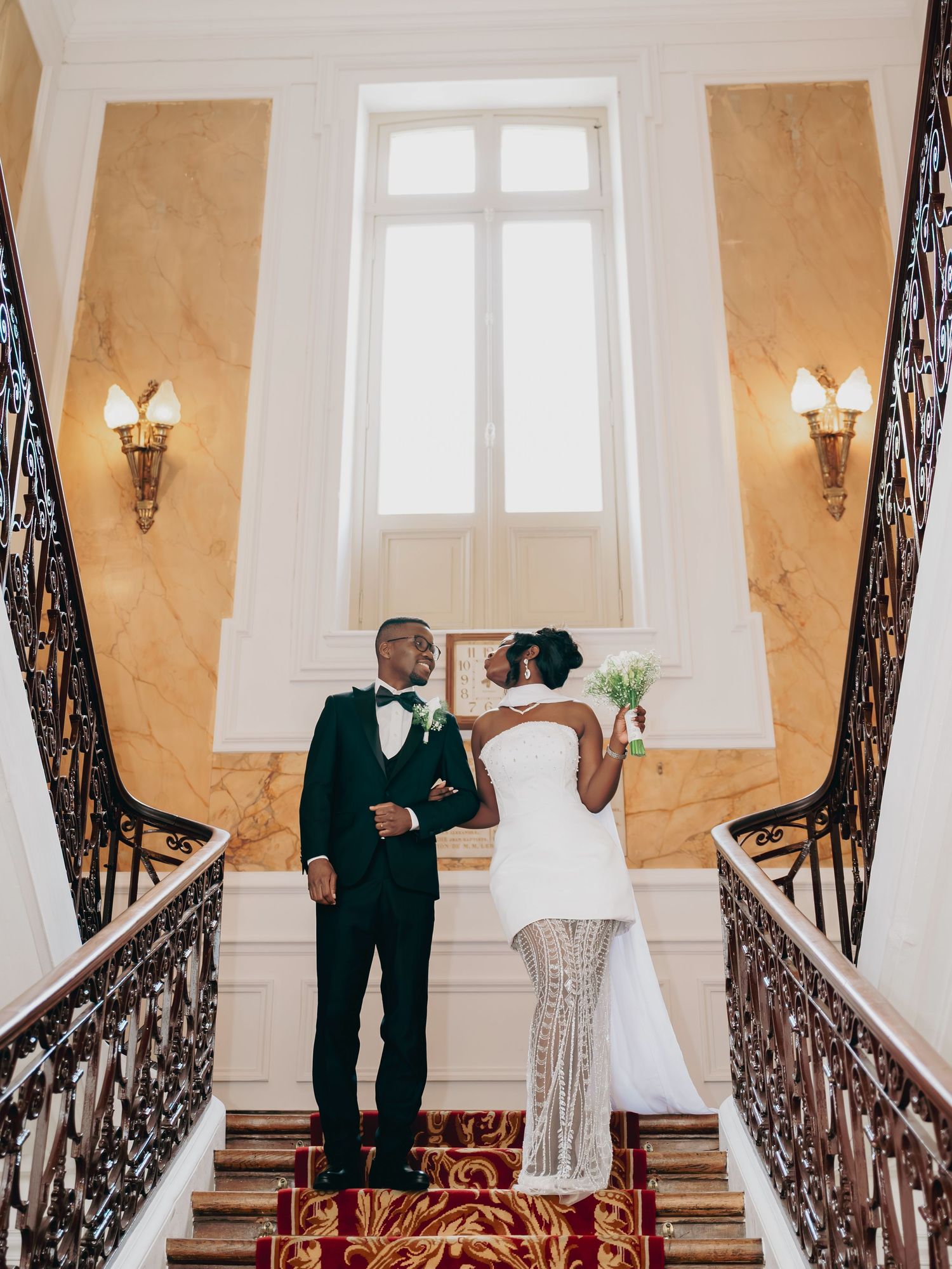 Bride and groom posing on grand staircase with ornate iron railings in elegant wedding venue.