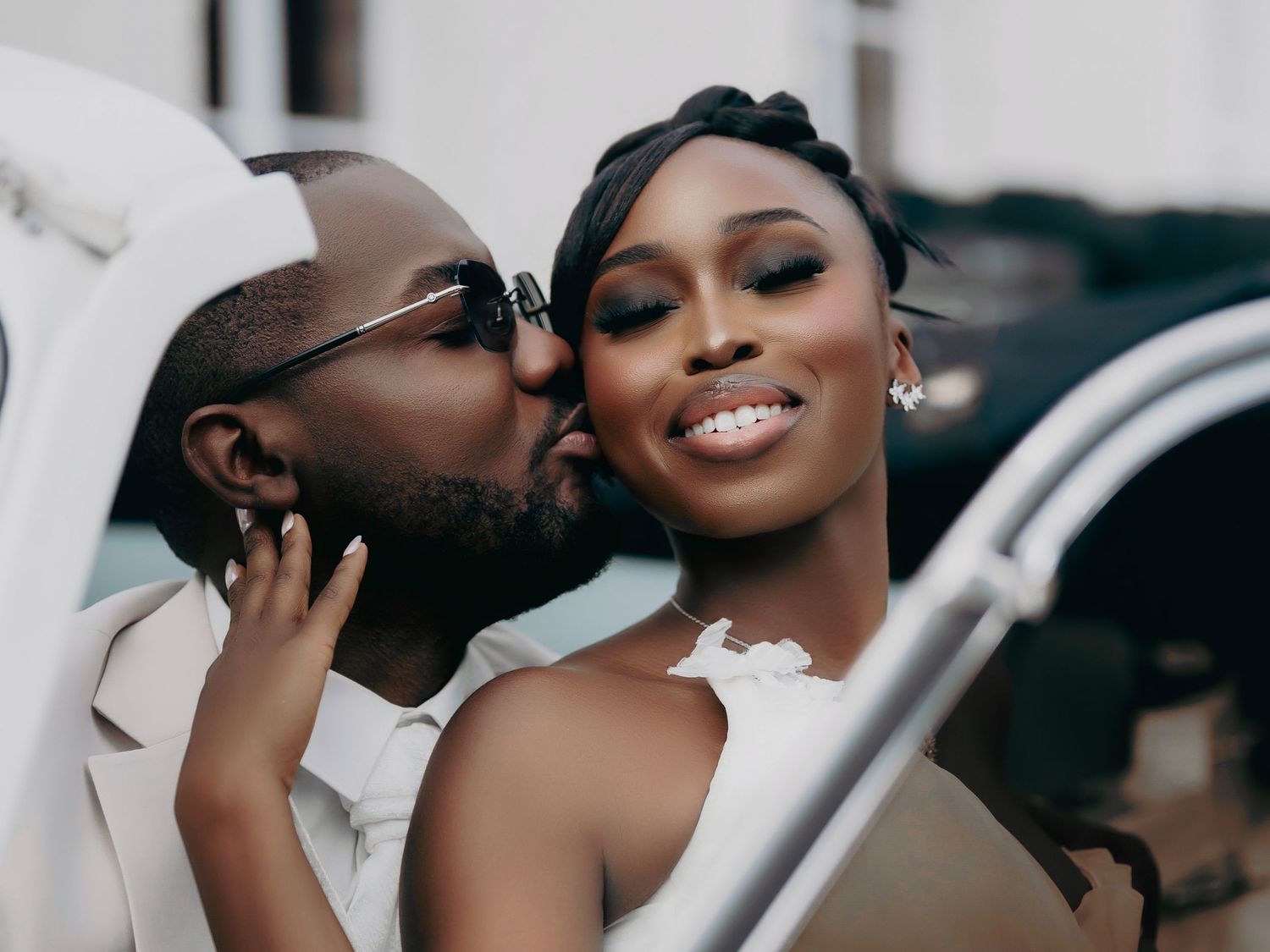 Groom kisses smiling brides cheek near a white vintage car on their wedding day.