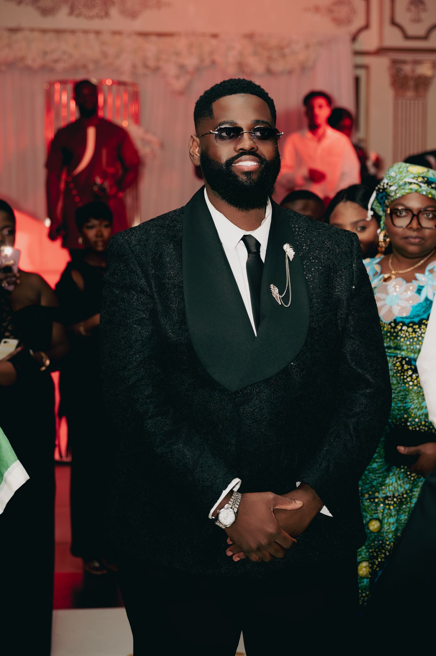 Groom in black tuxedo with sunglasses smiling at wedding reception.