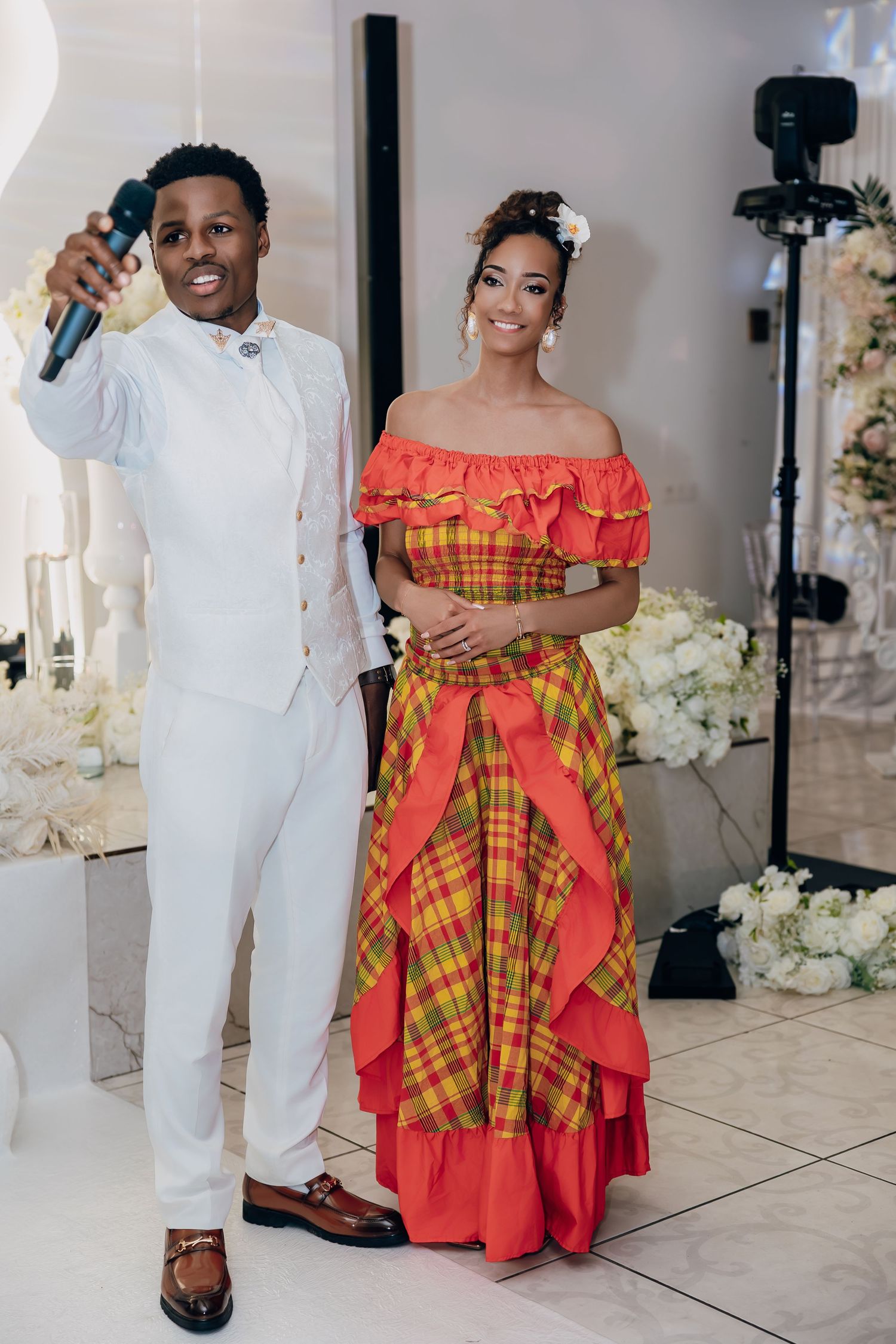 Bride in vibrant red and plaid Caribbean traditional dress with groom in white suit holding microphone at wedding.