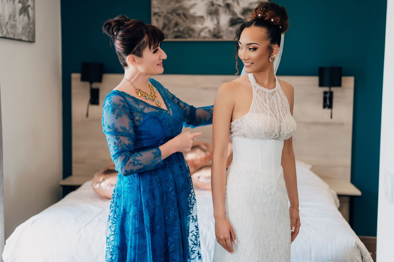 Mother in blue lace dress helps bride in halter neck wedding gown get ready in a teal-accented room.