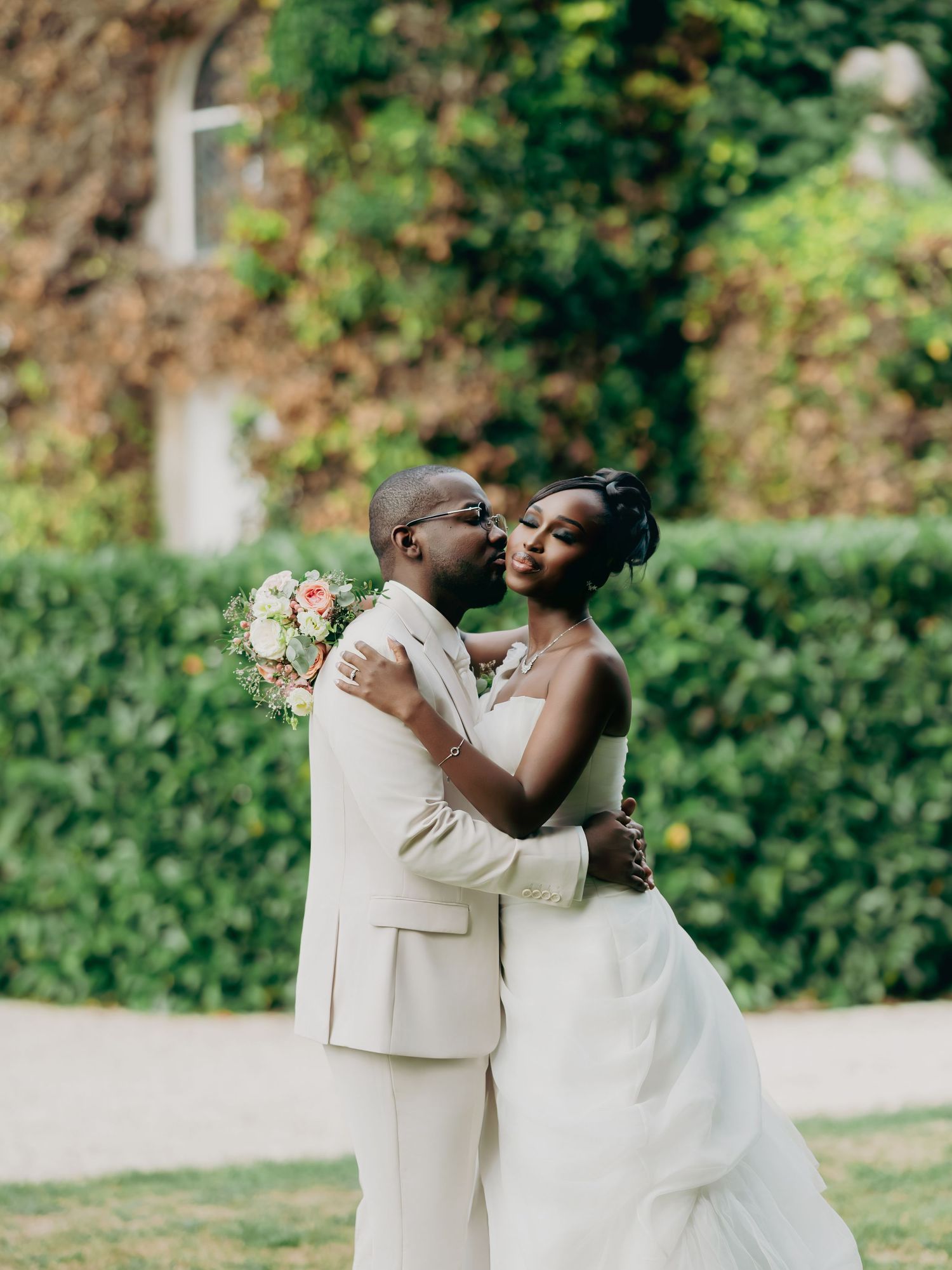Black newlyweds embrace and kiss in front of an ivy-covered building, bride holding pink bouquet.