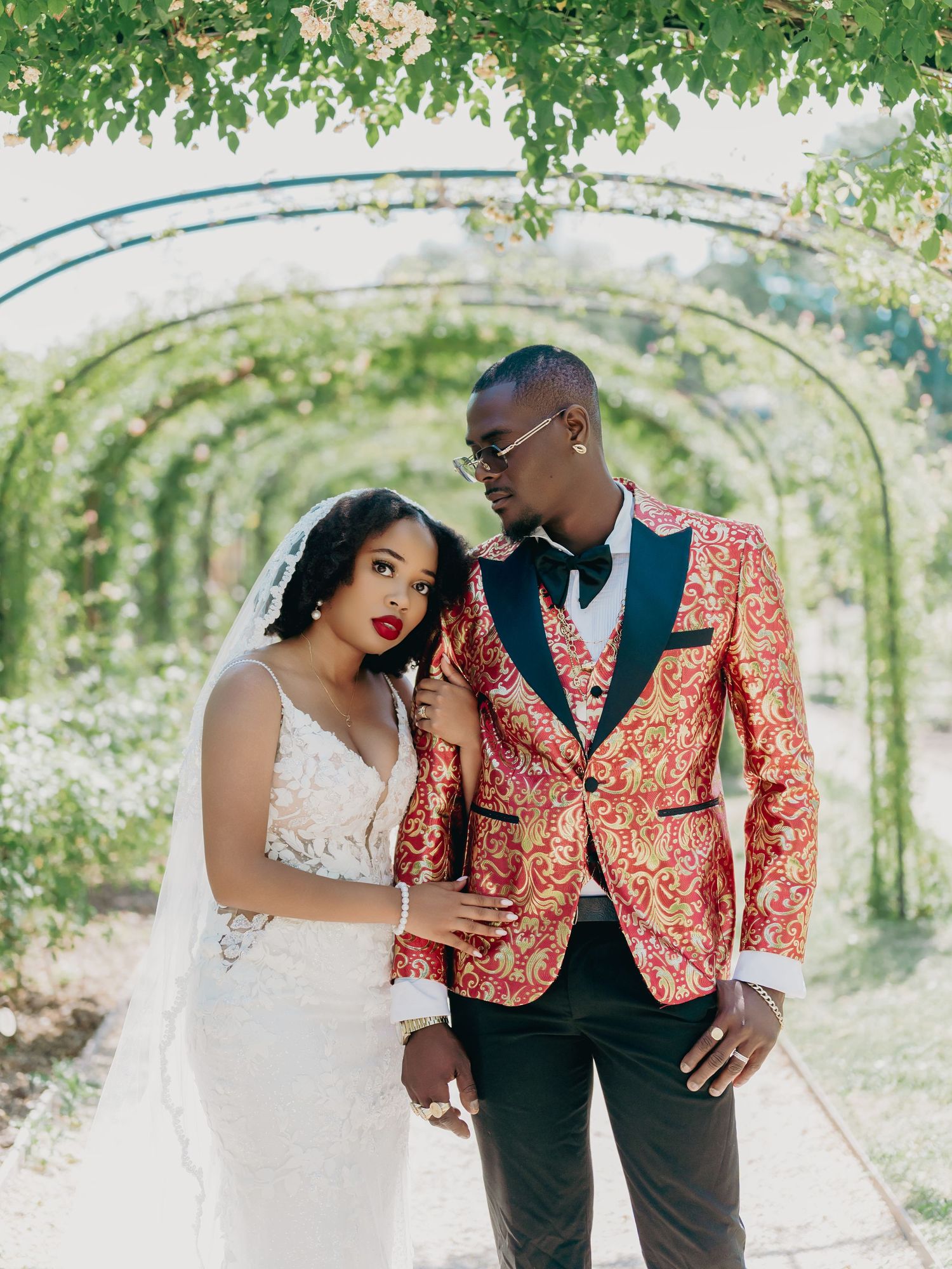 Wedding couple poses under green vine arch, groom in red gold blazer and bride in lace gown with veil.