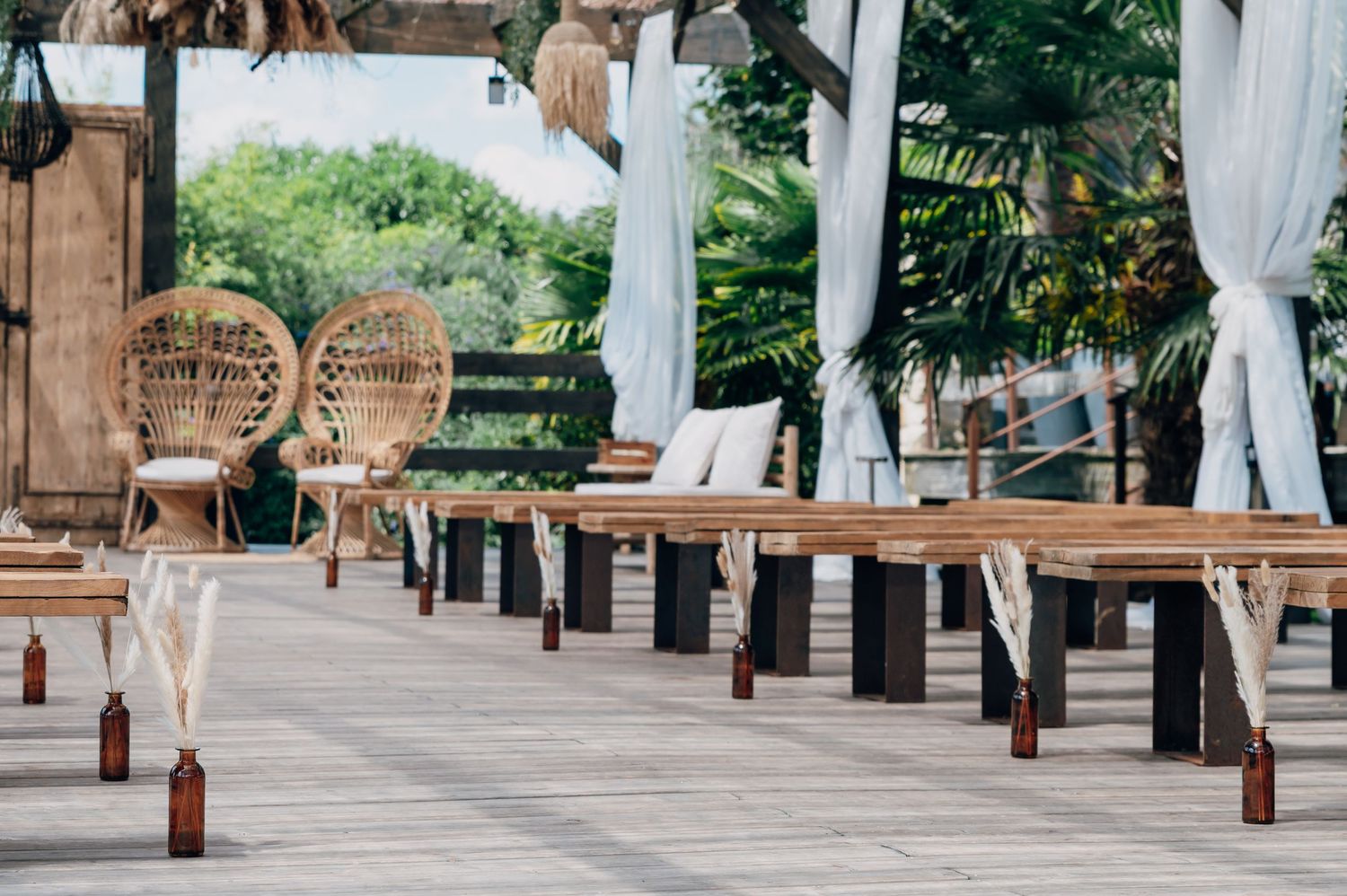Elegant outdoor wedding venue with wooden tables, rattan chairs, white drapes, and tropical greenery.