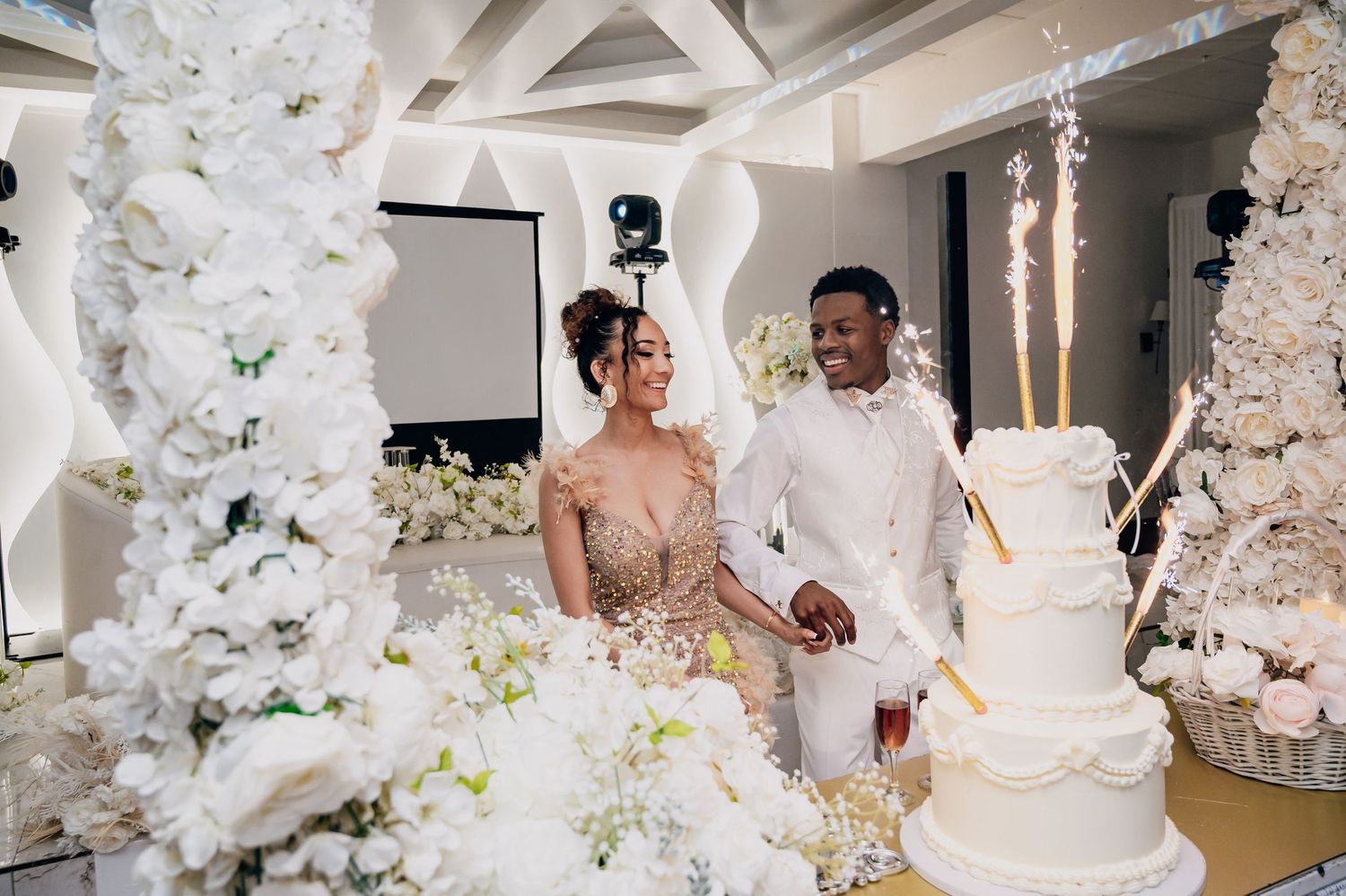 Bride and groom cut white wedding cake surrounded by white floral arrangements and sparklers.
